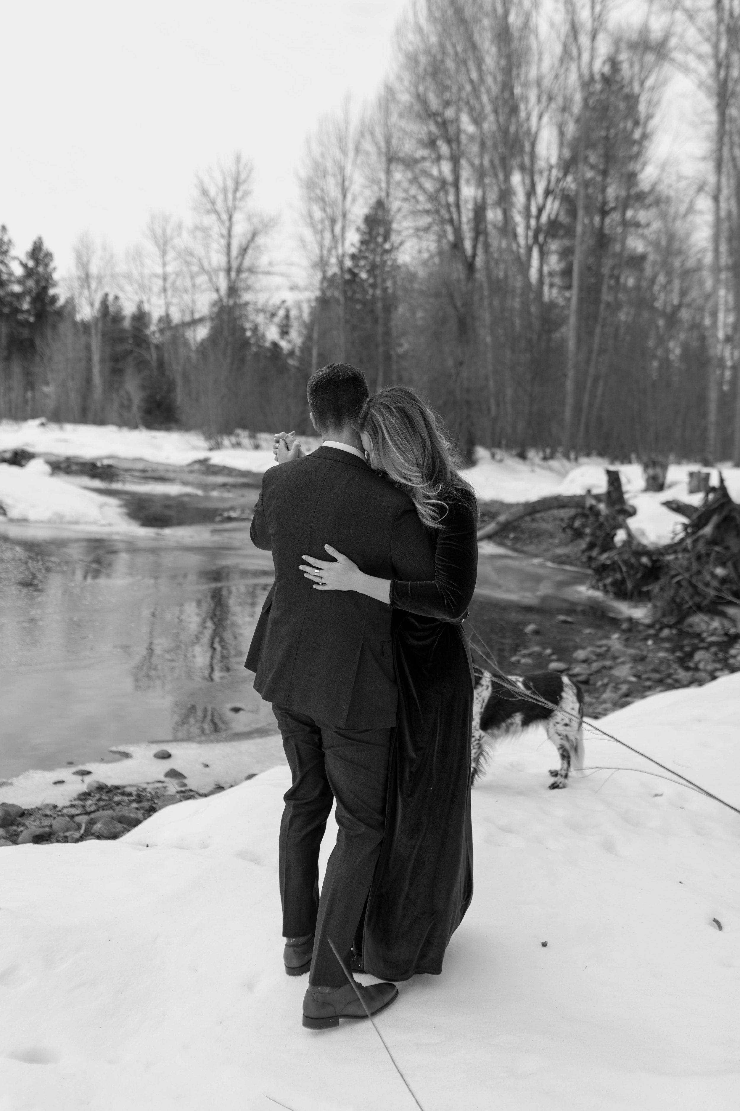 A couple embracing near a snowy riverbank, with a dog nearby. The woman wears a long dress and the man is in a suit, set in a wooded, winter landscape.