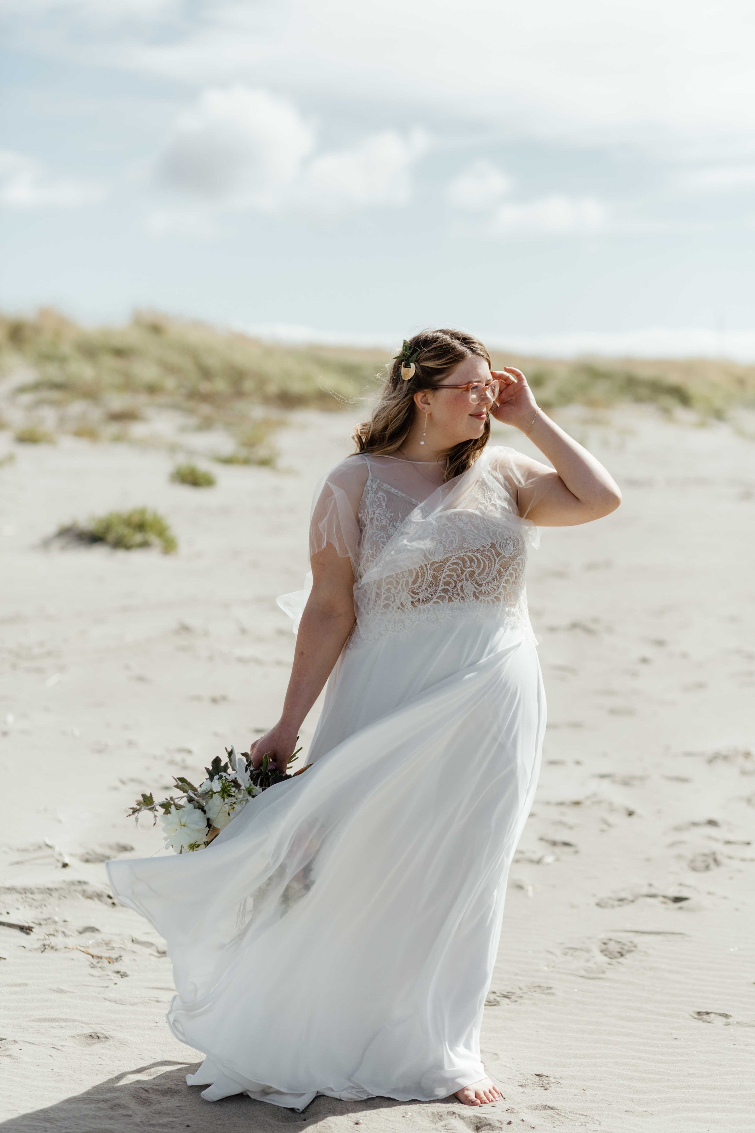 Woman in a white wedding dress standing on a sandy beach, holding a bouquet of flowers, under a clear sky.