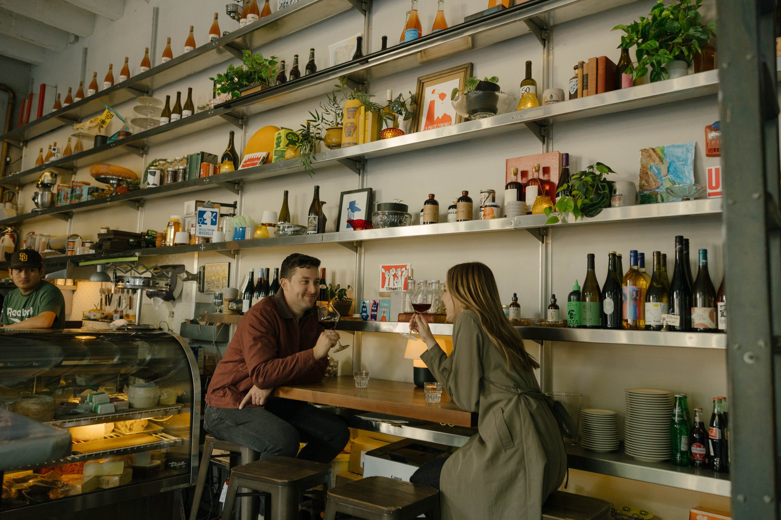 Cozy cafe interior with two people enjoying drinks at a wooden table, surrounded by shelves filled with bottles, plants, and various items. A staff member works near a refrigerated display with pastries.