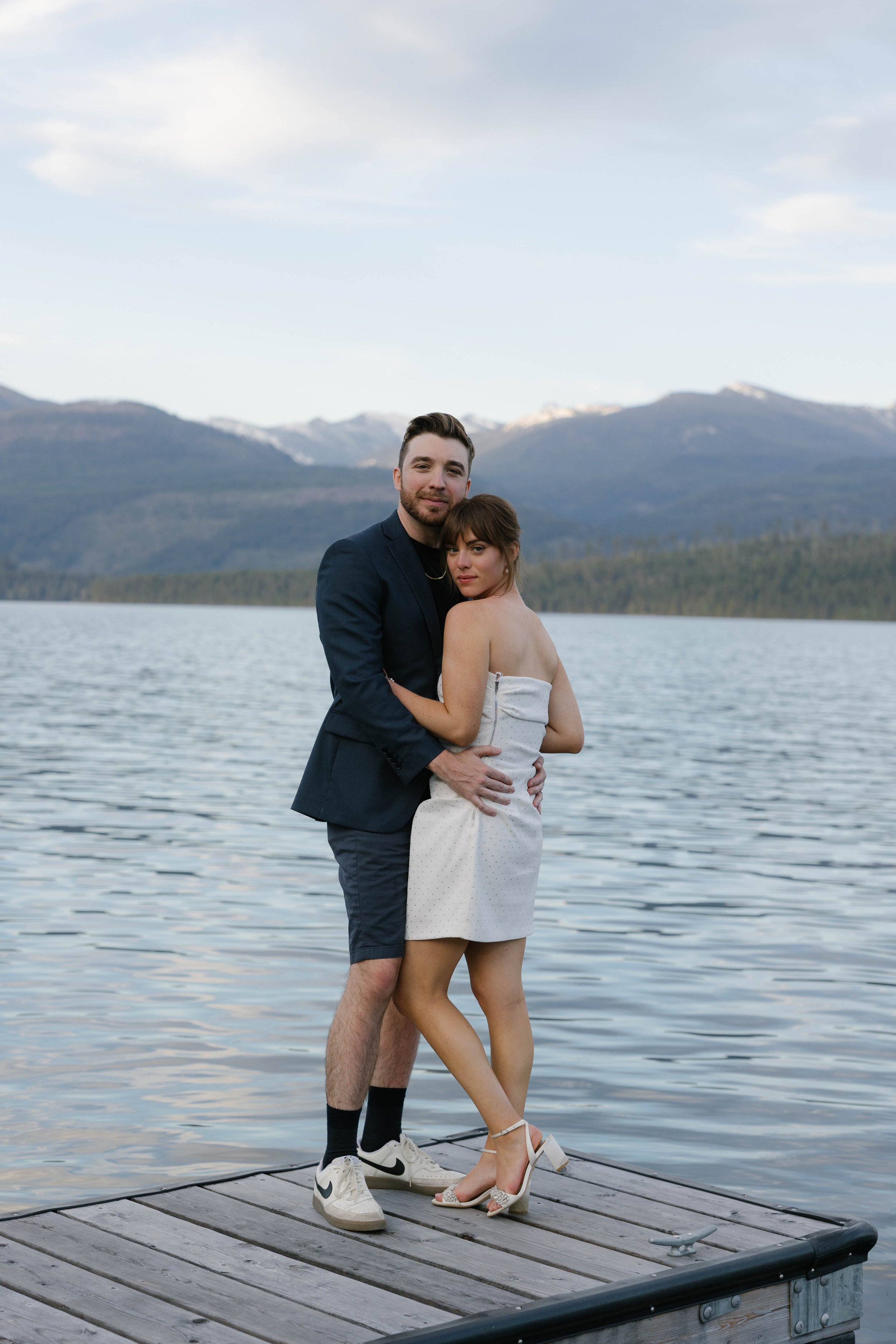 A couple embracing on a wooden dock by a lake with mountains in the background.