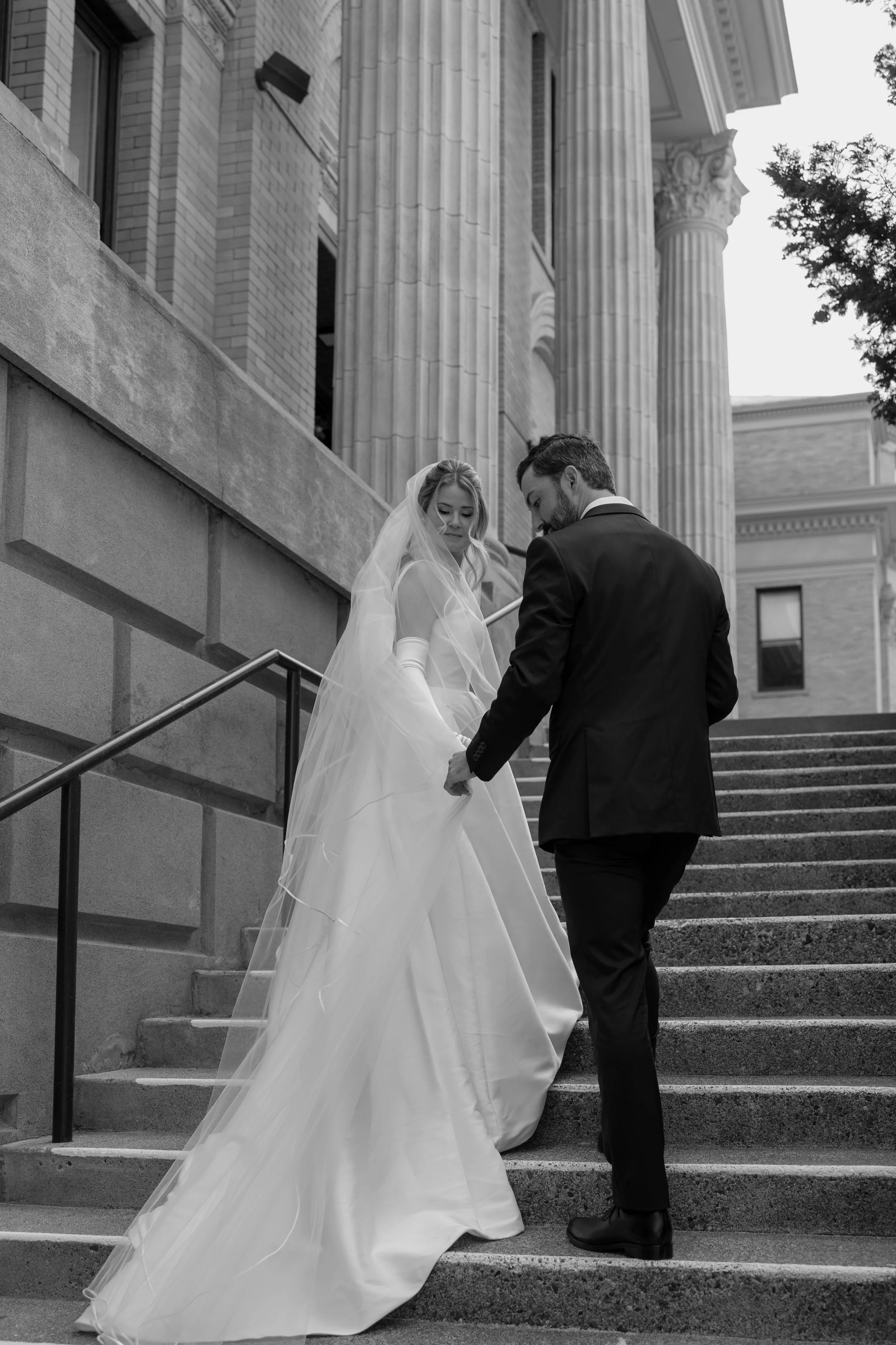 Bride and groom walking up stairs, bride in white dress with veil, groom in suit, historic building with columns in background.