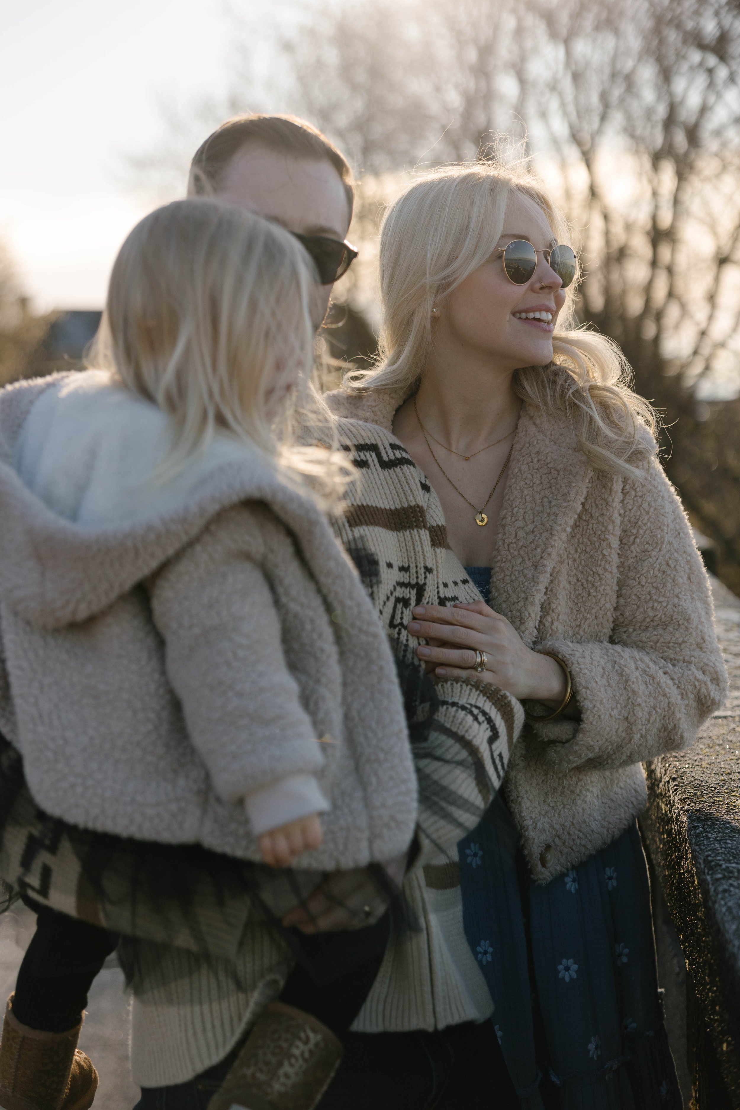 A family of three, including a woman, a man, and a child, wearing winter coats and sunglasses, standing outdoors on a sunny day with bare trees in the background.
