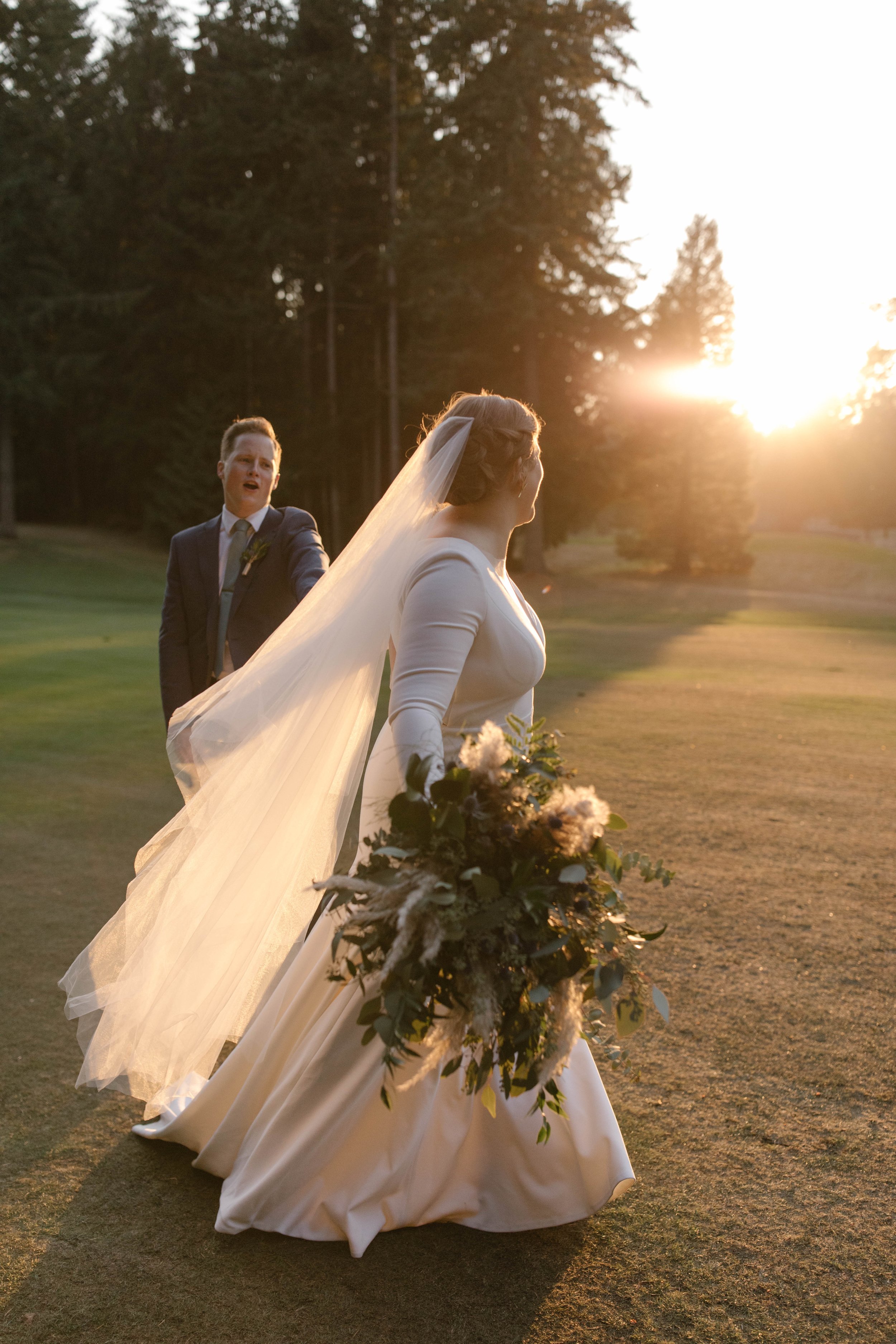 Bride and groom walking in a sunlit field, bride holding a bouquet, veil flowing, forest in the background.
