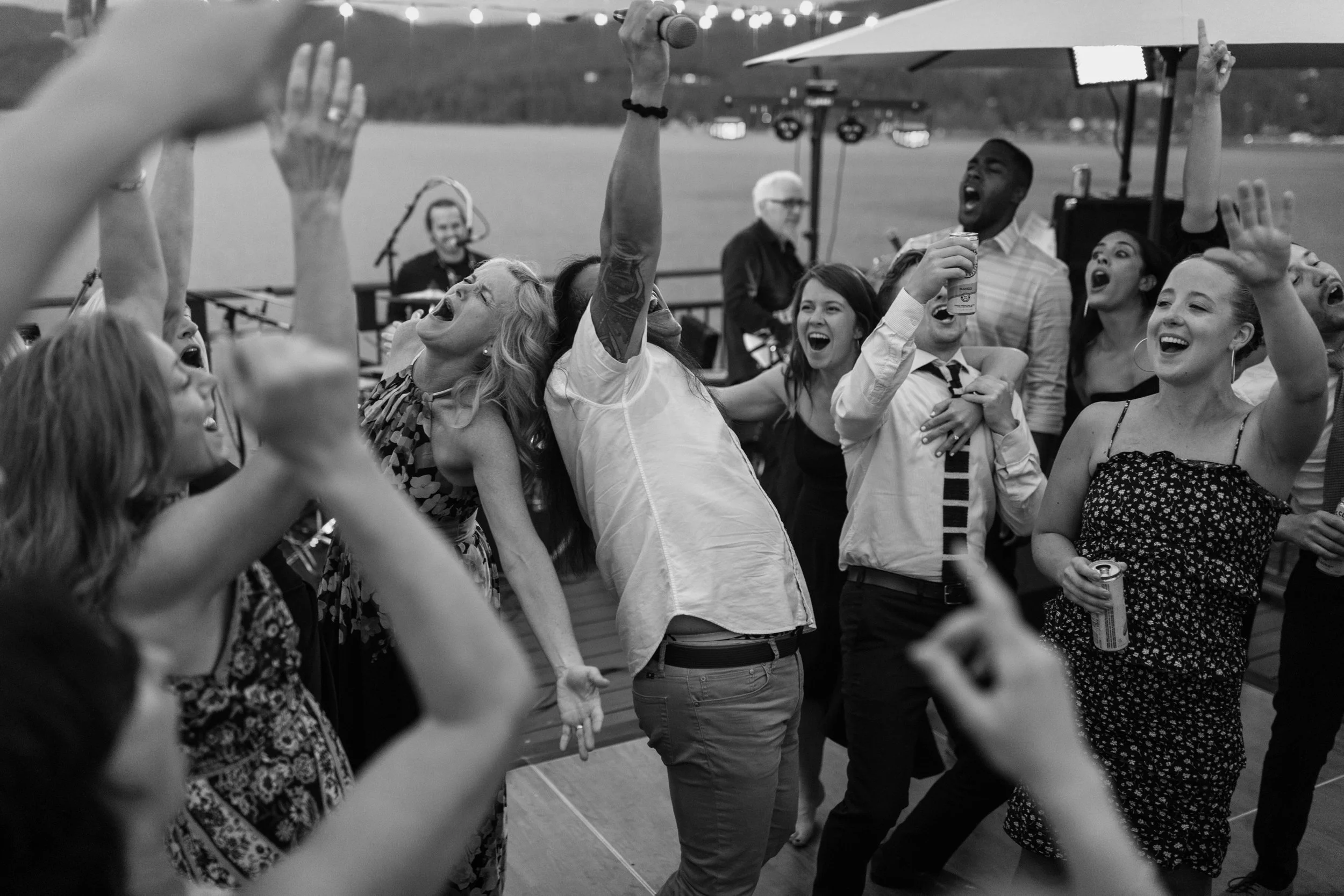 Group of people energetically dancing and cheering at an outdoor event, holding drinks, with a background of a lake and string lights.
