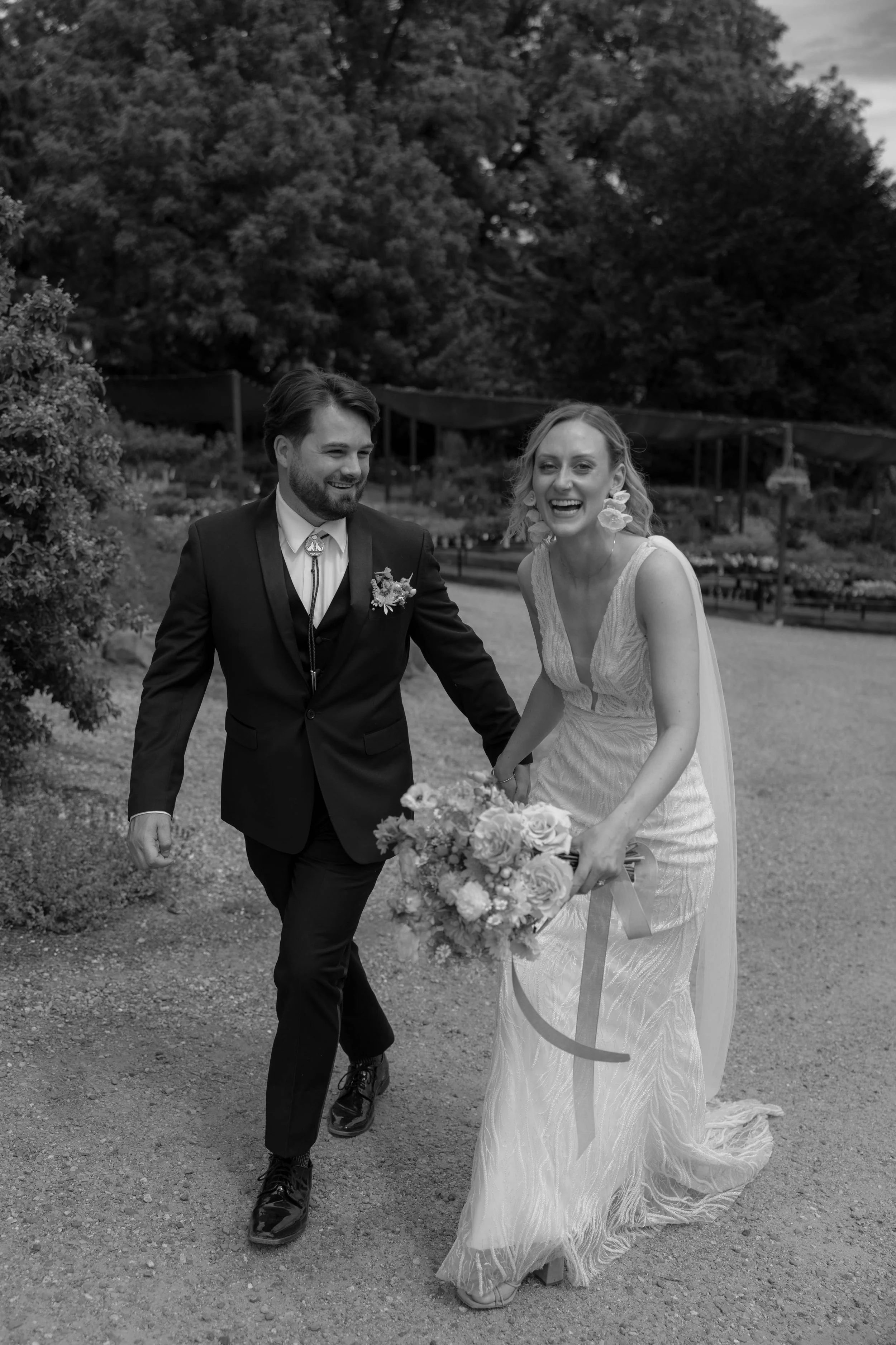 Bride and groom laughing and walking outdoors, bride holding bouquet, in black and white.