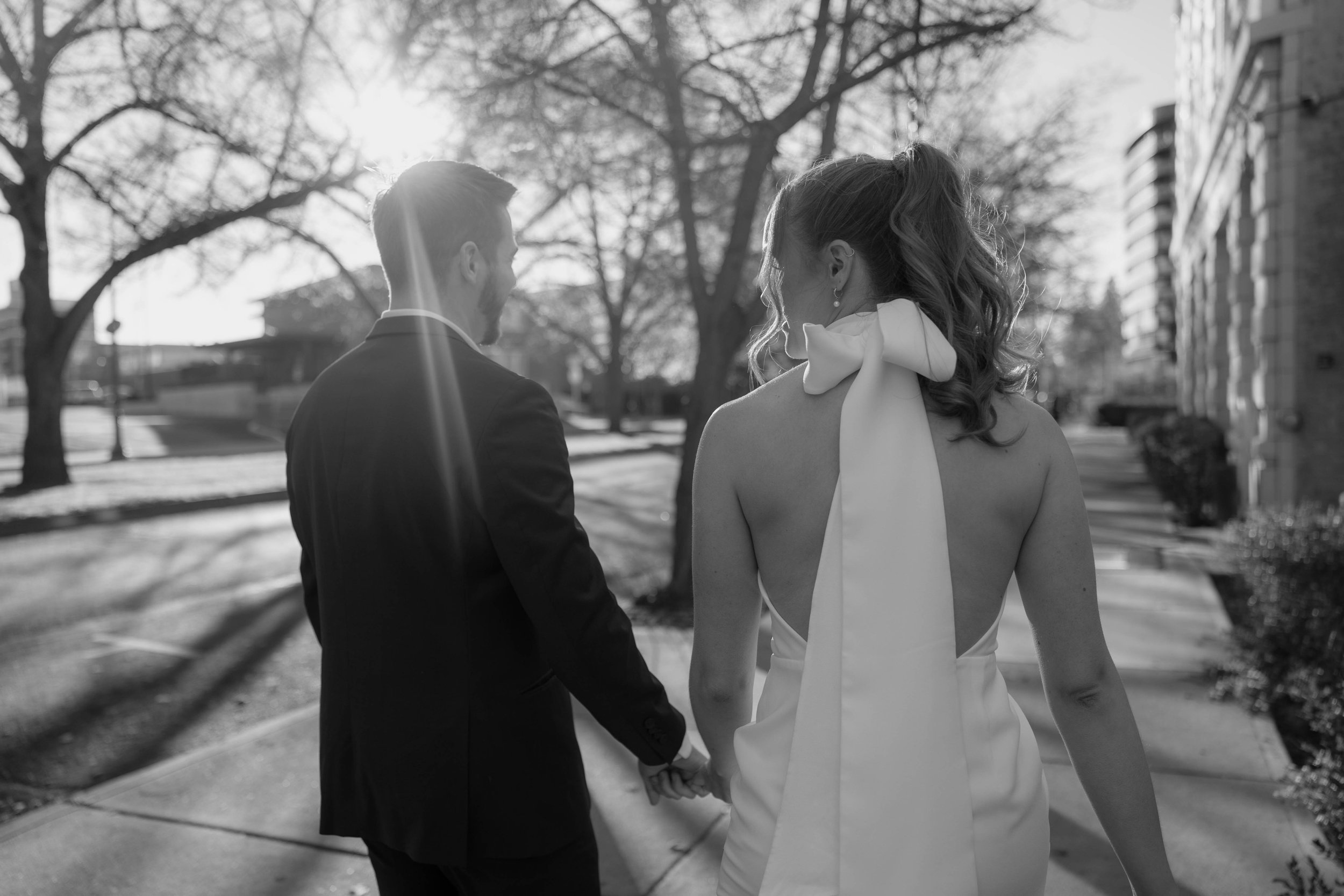 Black and white photo of a couple walking hand in hand, dressed formally, with sunlight filtering through trees.