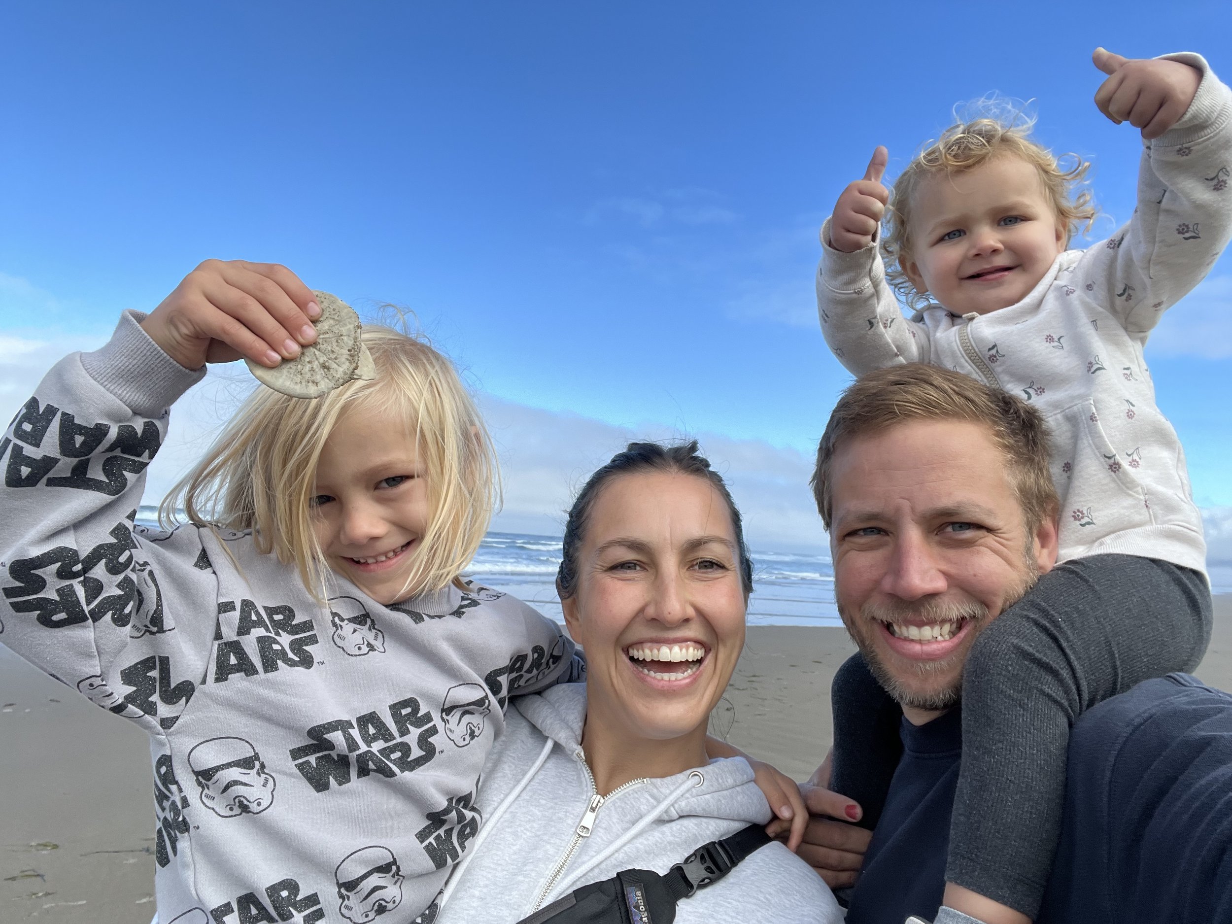 A family of four smiling at a beach, with one child holding a sand dollar and the other giving thumbs up.
