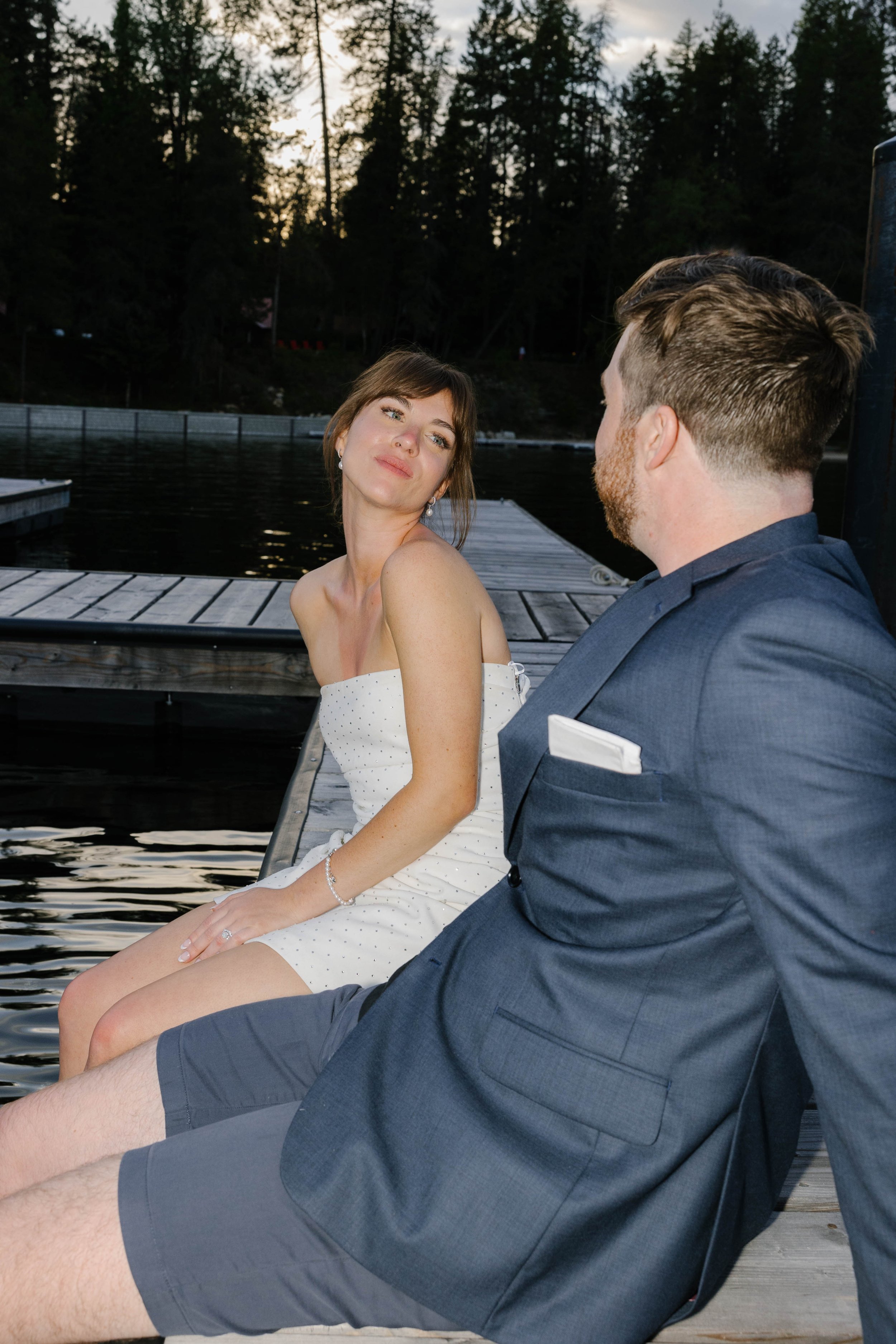 A woman in a white dress and a man in a suit sitting on a dock by a lake, surrounded by trees, during sunset.