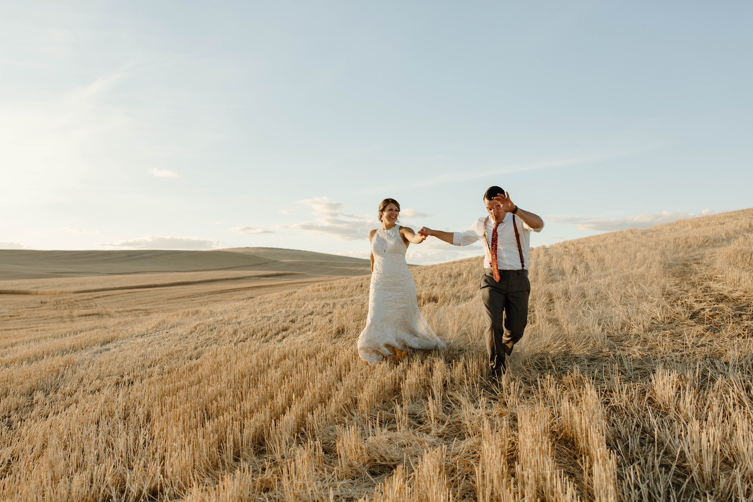 Bride and groom holding hands, walking through a field of dry grass, dressed in wedding attire, under a clear sky.