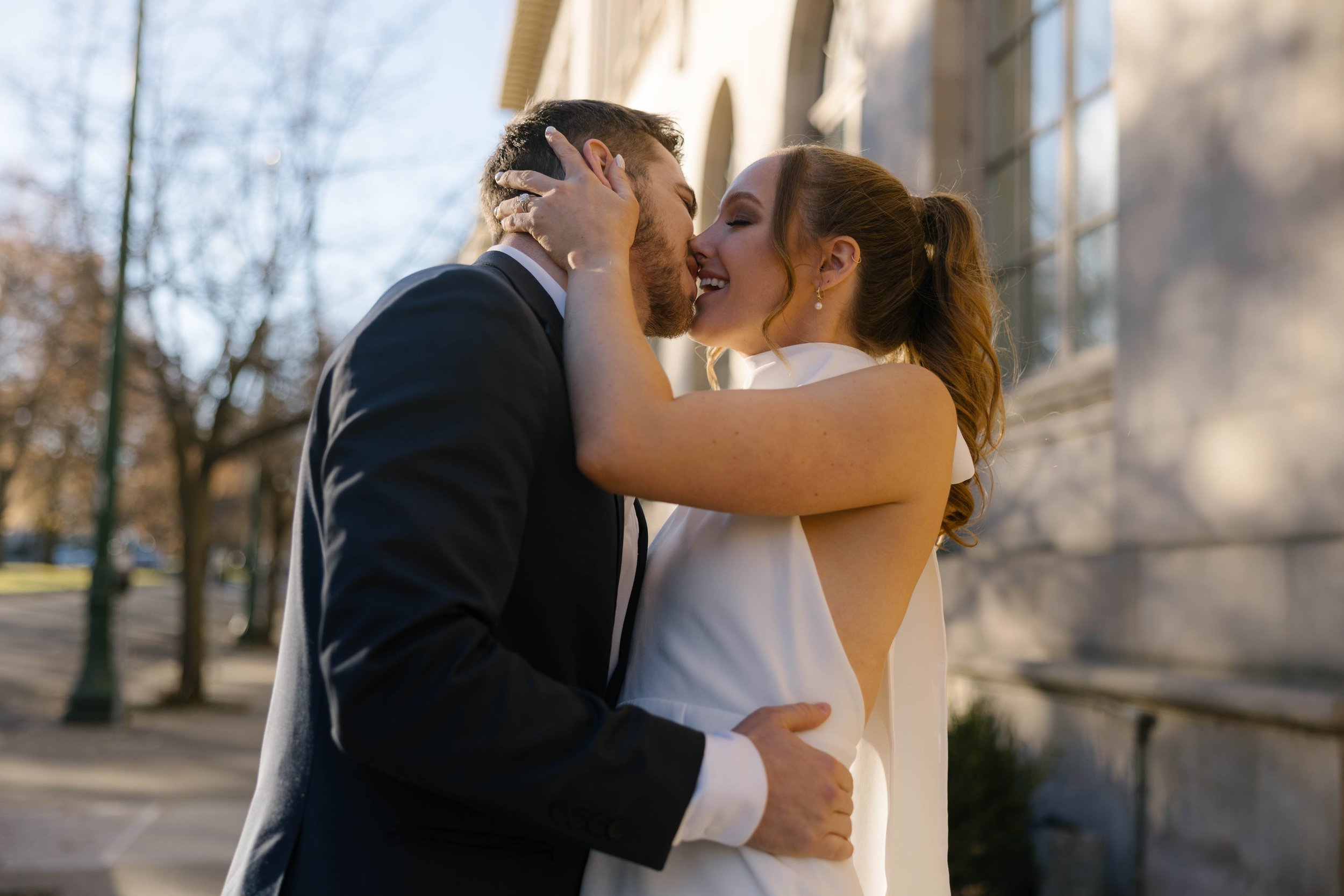 A couple embracing and smiling outdoors in formal attire, with trees and a building in the background.
