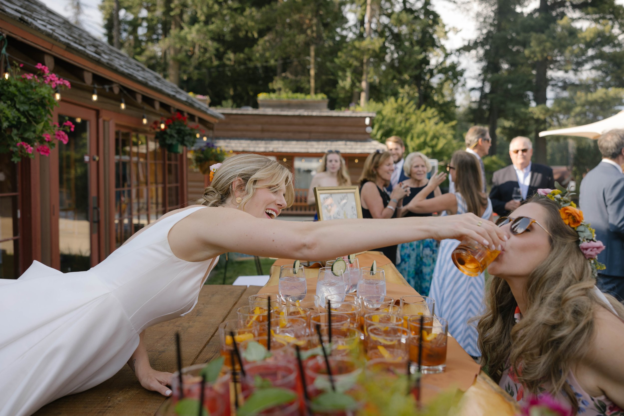 Woman in white dress playfully feeds a drink to another woman wearing sunglasses and a flower crown at an outdoor event, with guests in the background.