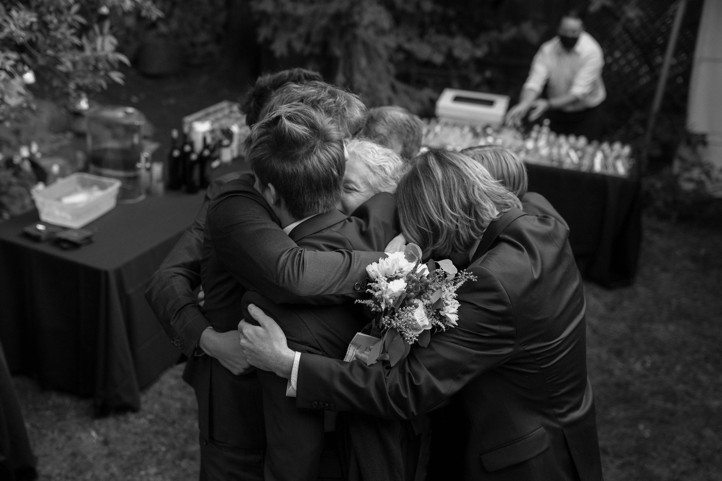 Group hug at an outdoor event, people wearing formal attire, with a floral bouquet visible in one person's hand.