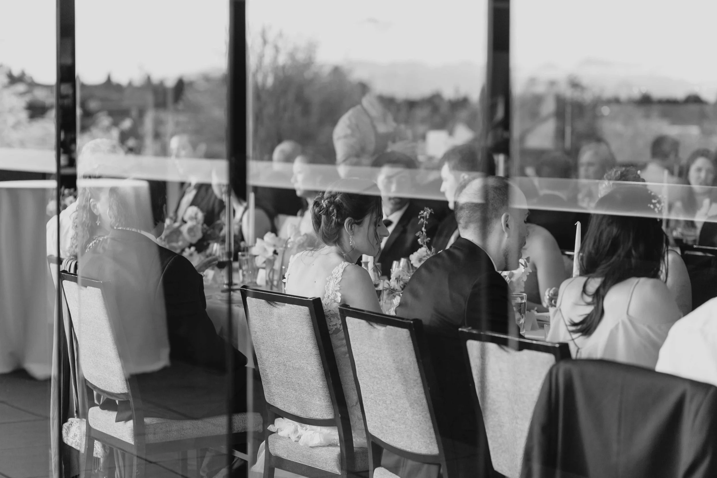 Black and white photo of wedding guests seated at a reception table, visible through a glass window, reflecting the outdoor scenery.