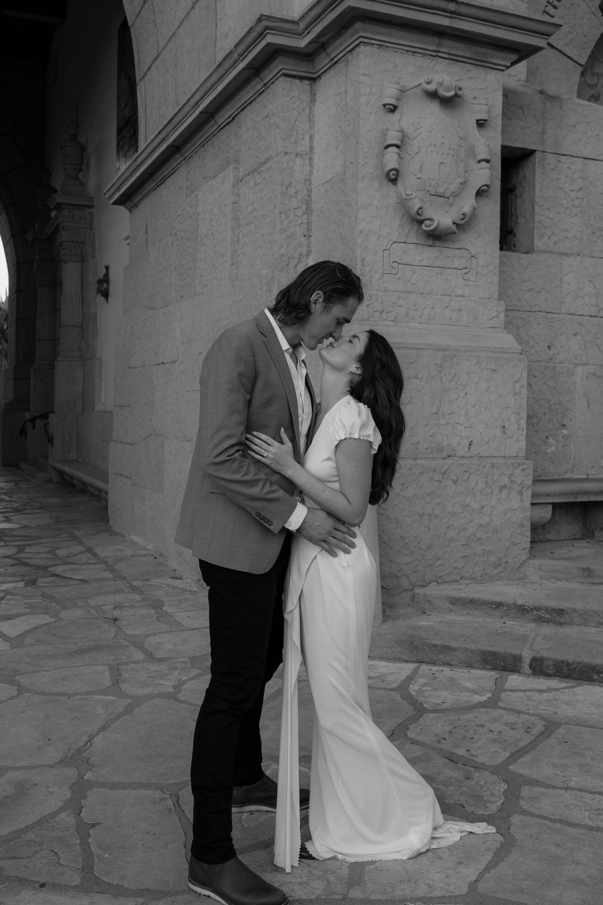 A couple embracing and kissing in front of a stone building, with the woman in a long white dress and the man in a suit, black and white image.