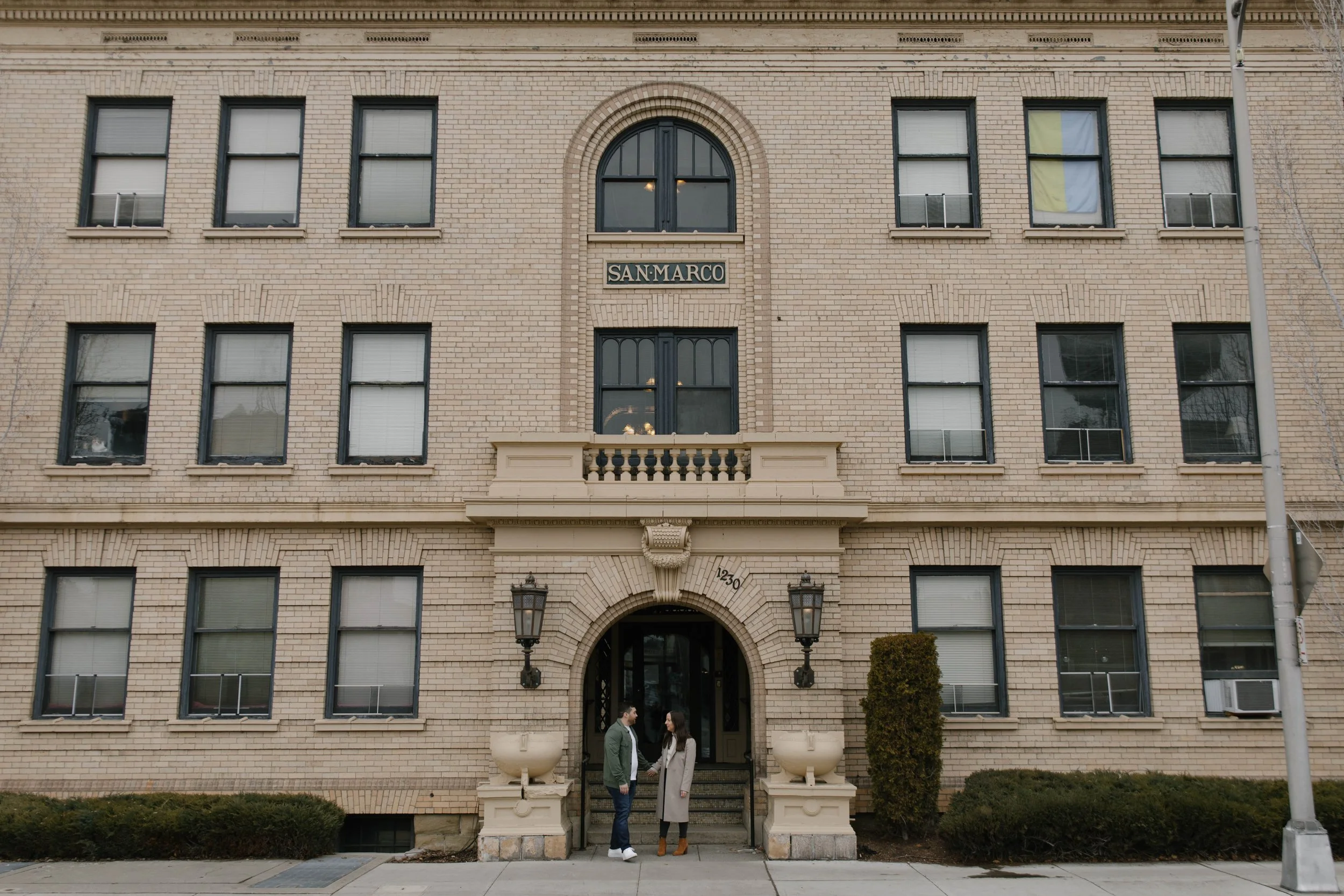 Front view of a three-story brick building with black-framed windows and an arched entryway. The building has a sign "San Marco" above the entrance, two large lamps beside the entry, and two people standing near the doorway. Sidewalk and small shrubs