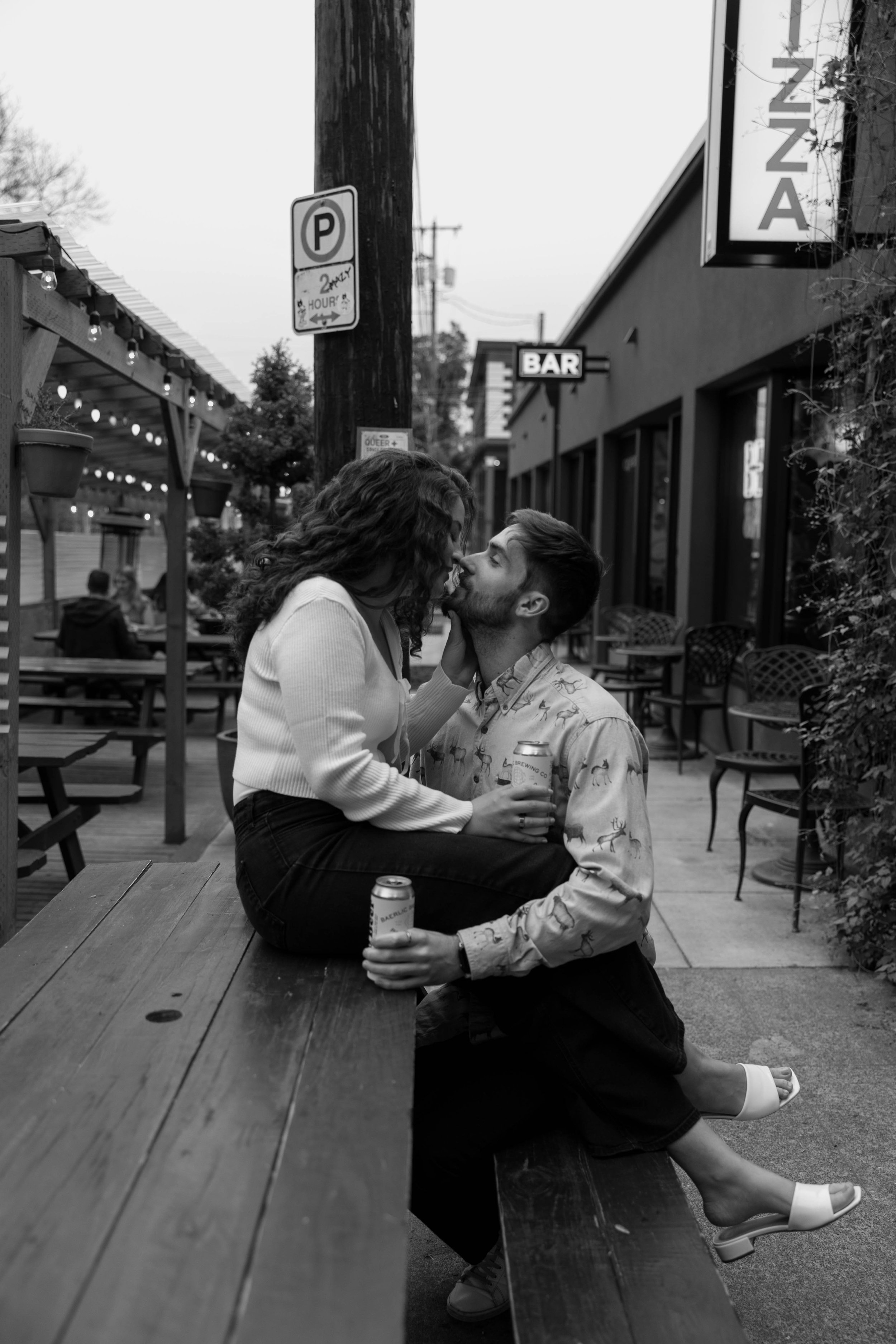 Black and white photo of a couple sitting on a wooden table outdoors, holding cans, and sharing a moment. "Pizza" and "Bar" signs visible in the background.