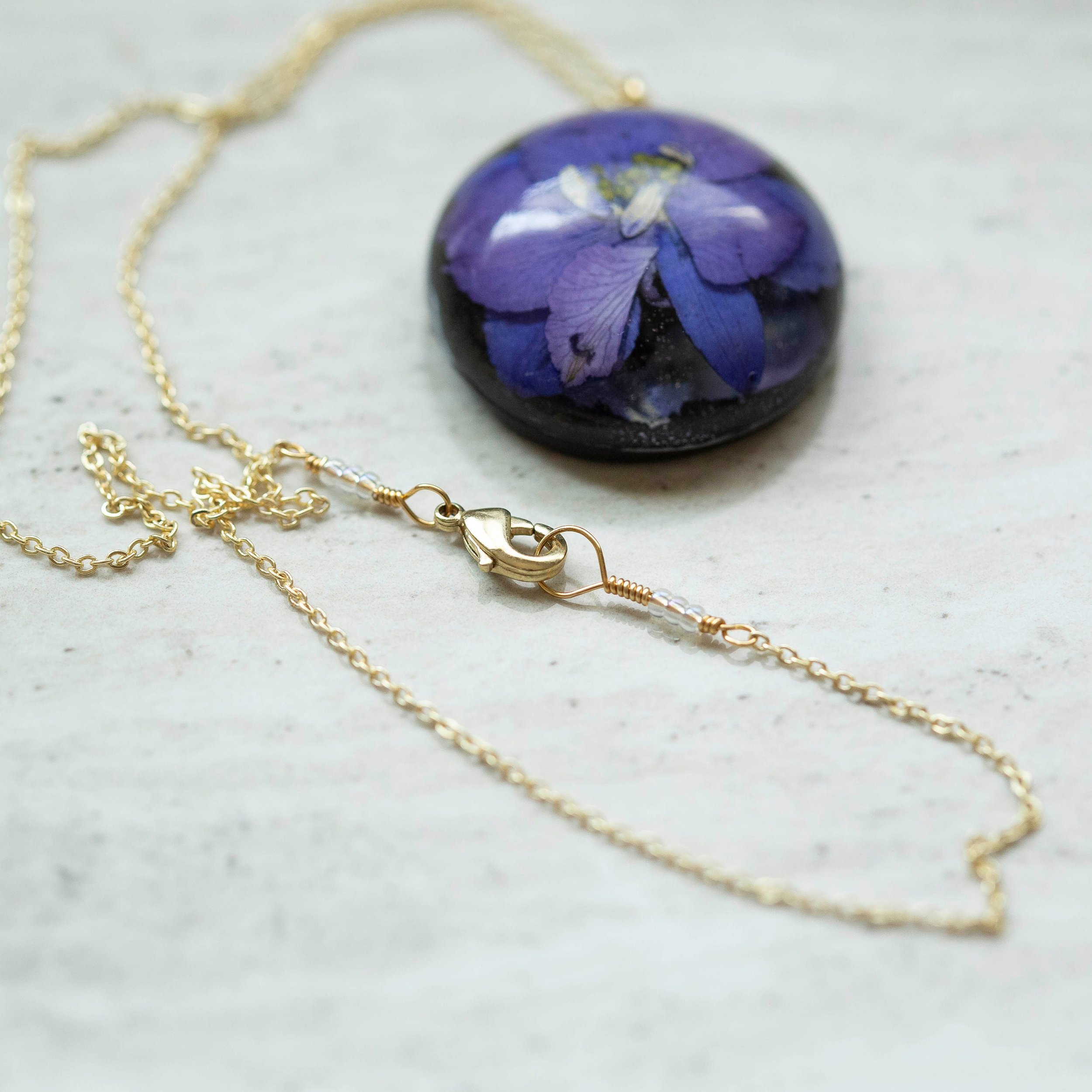 Beautiful real Purple Flower In a resin dome with a black clay back on a gold chain.