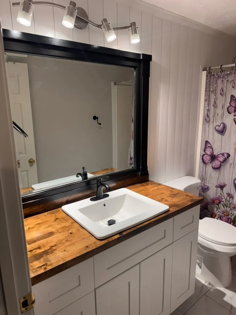 White shaker bathroom vanity with stained butcher block top and black framed mirror