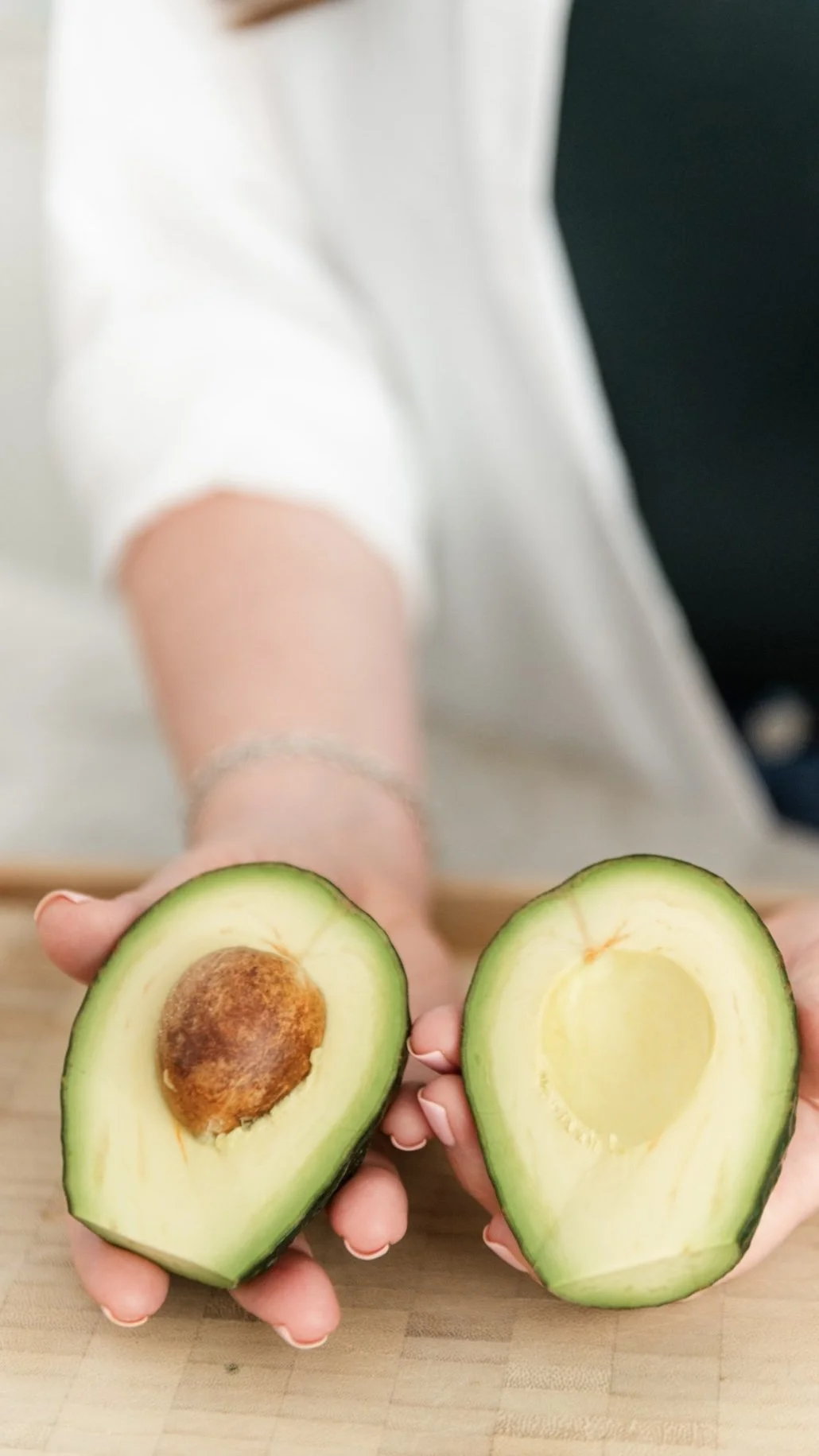 A person holding two halves of an avocado over a wooden surface, with the seed in one half and the empty cavity in the other half.