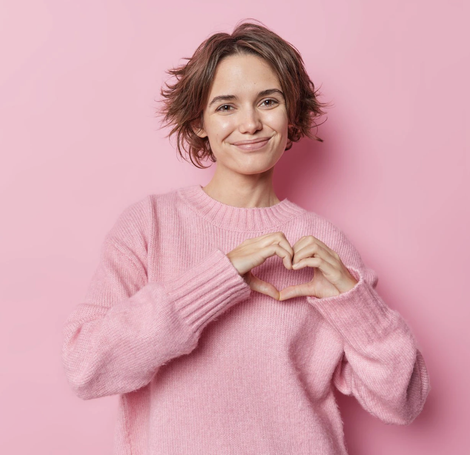 A young woman with short, wavy hair smiling at the camera, wearing a pink sweater, and making a heart shape with her hands against a pink background.
