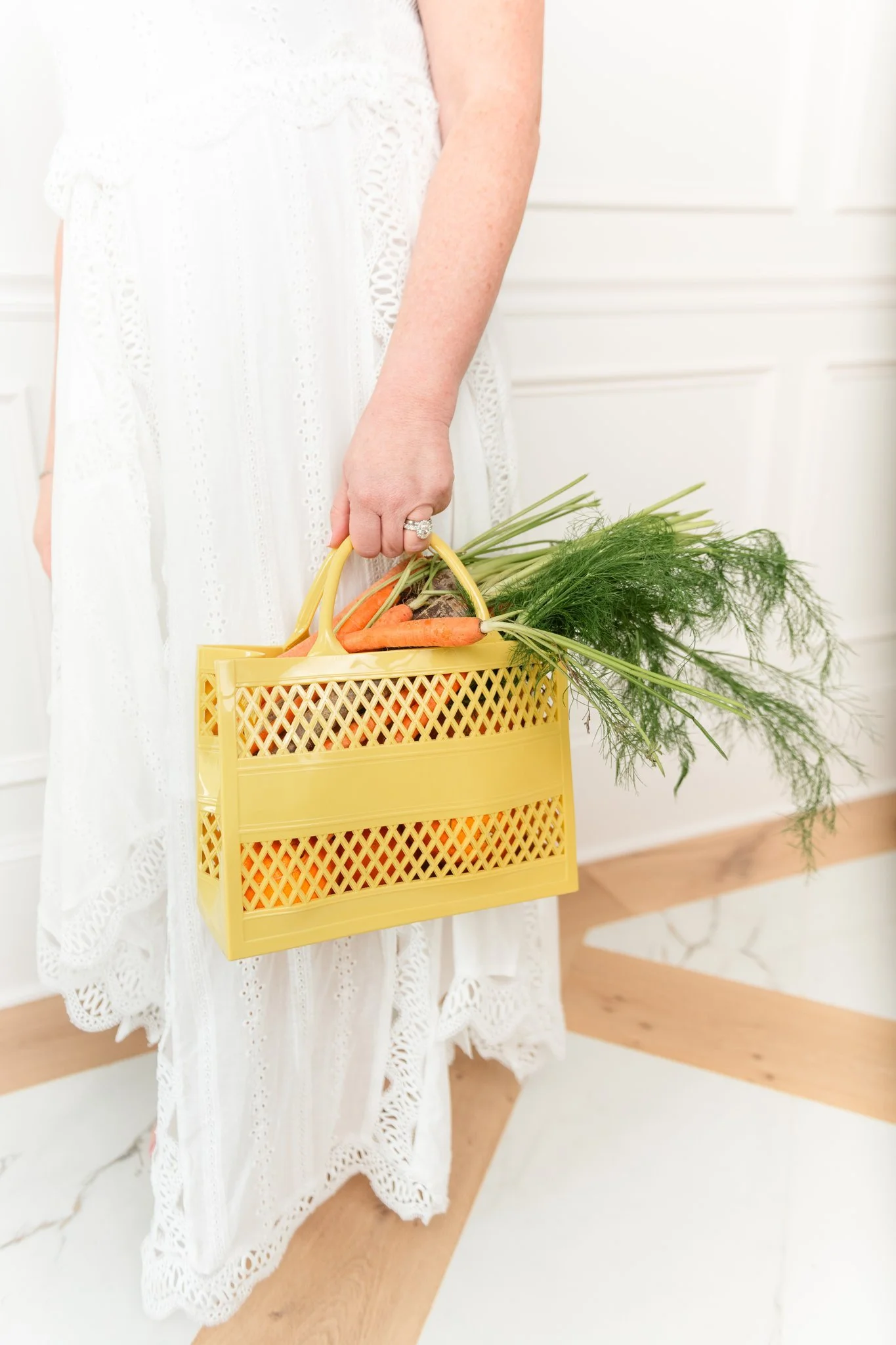 Person wearing a white dress holding a yellow basket with carrots and greens.