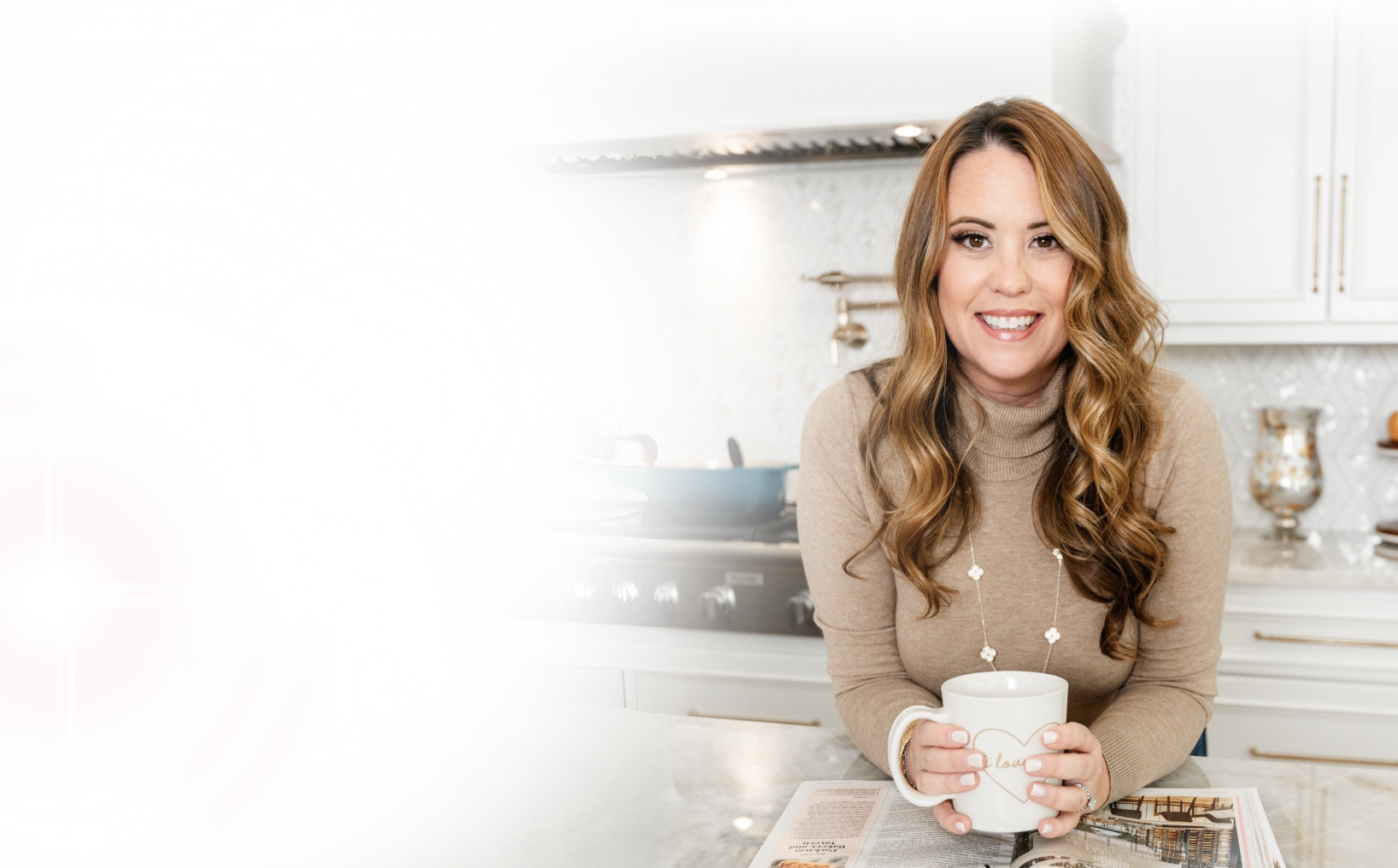 A woman with long wavy hair smiling and holding a coffee mug in a bright, modern kitchen