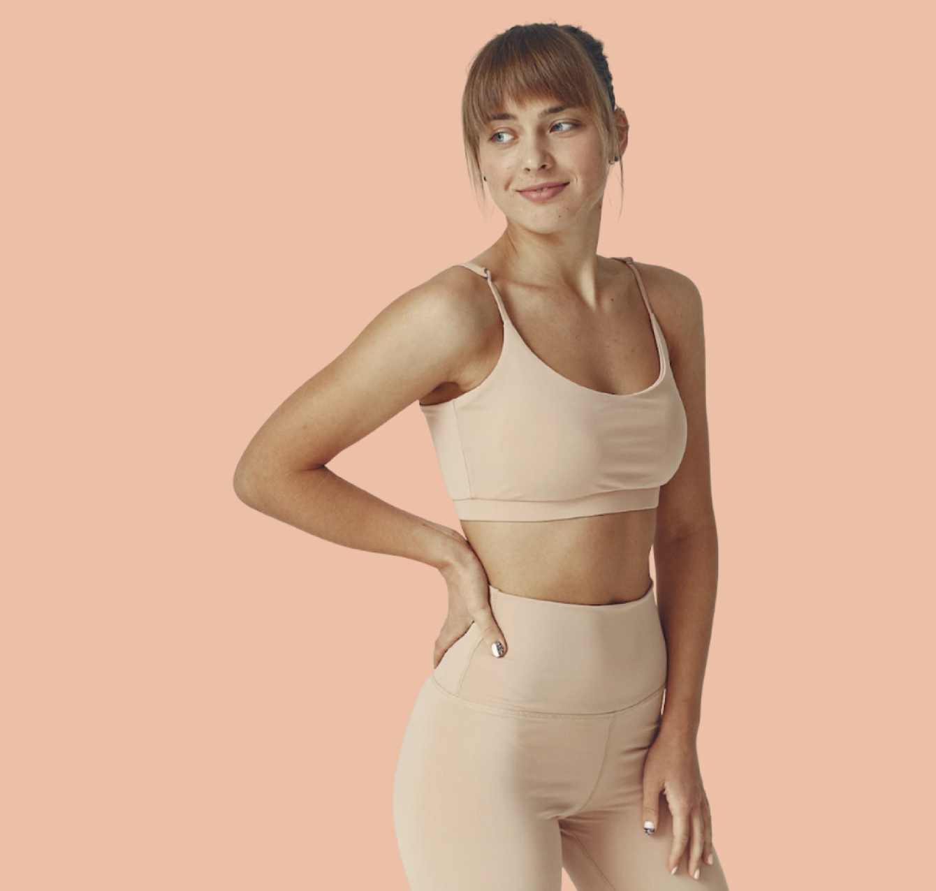 A woman in a beige sports bra and matching high-waisted leggings standing against a peach background, smiling softly and looking to her left.