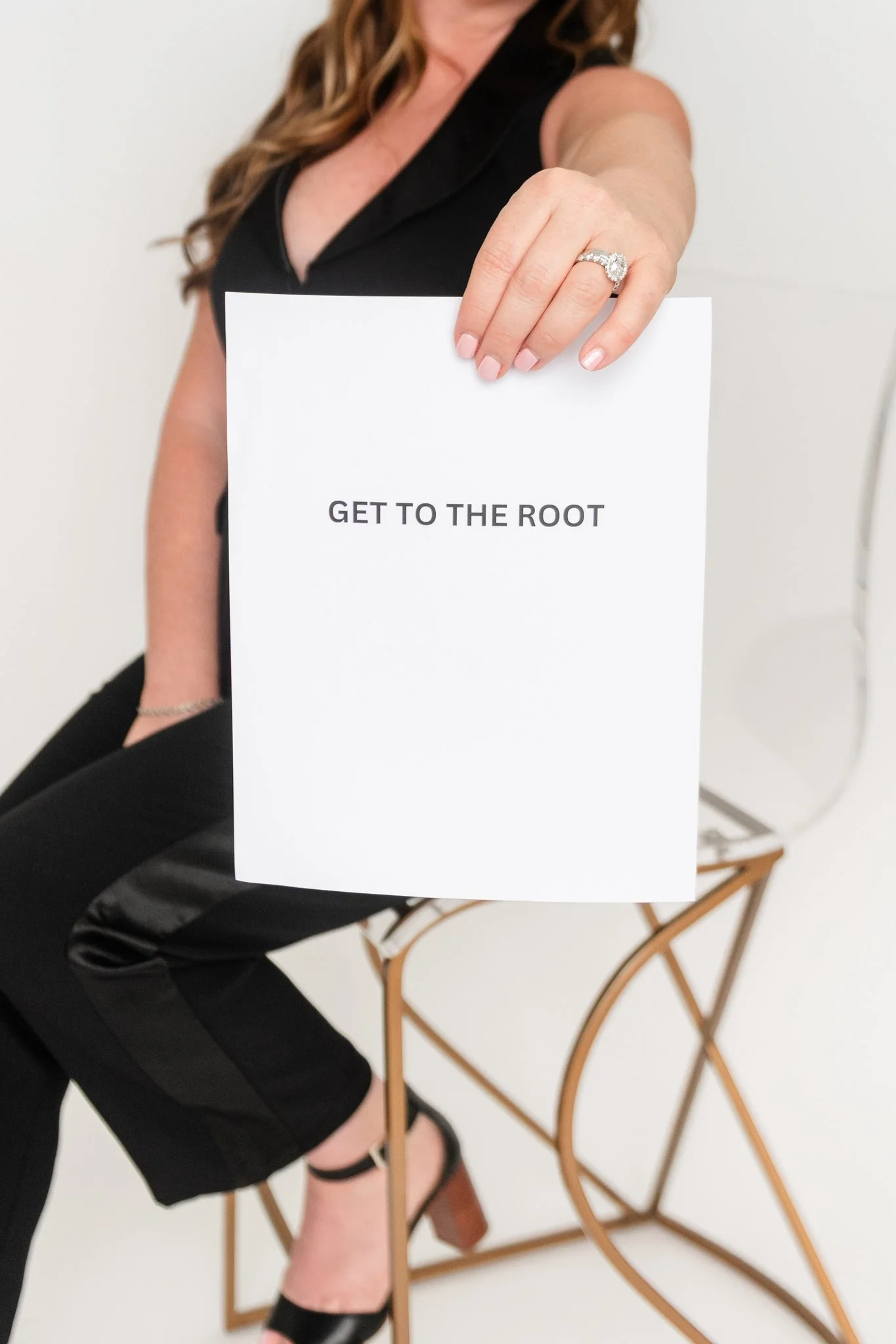 A woman sitting on a chair, holding a white sign with the text "GET TO THE ROOT." She is wearing a black sleeveless outfit and black high heels, and has a large diamond ring on her finger.