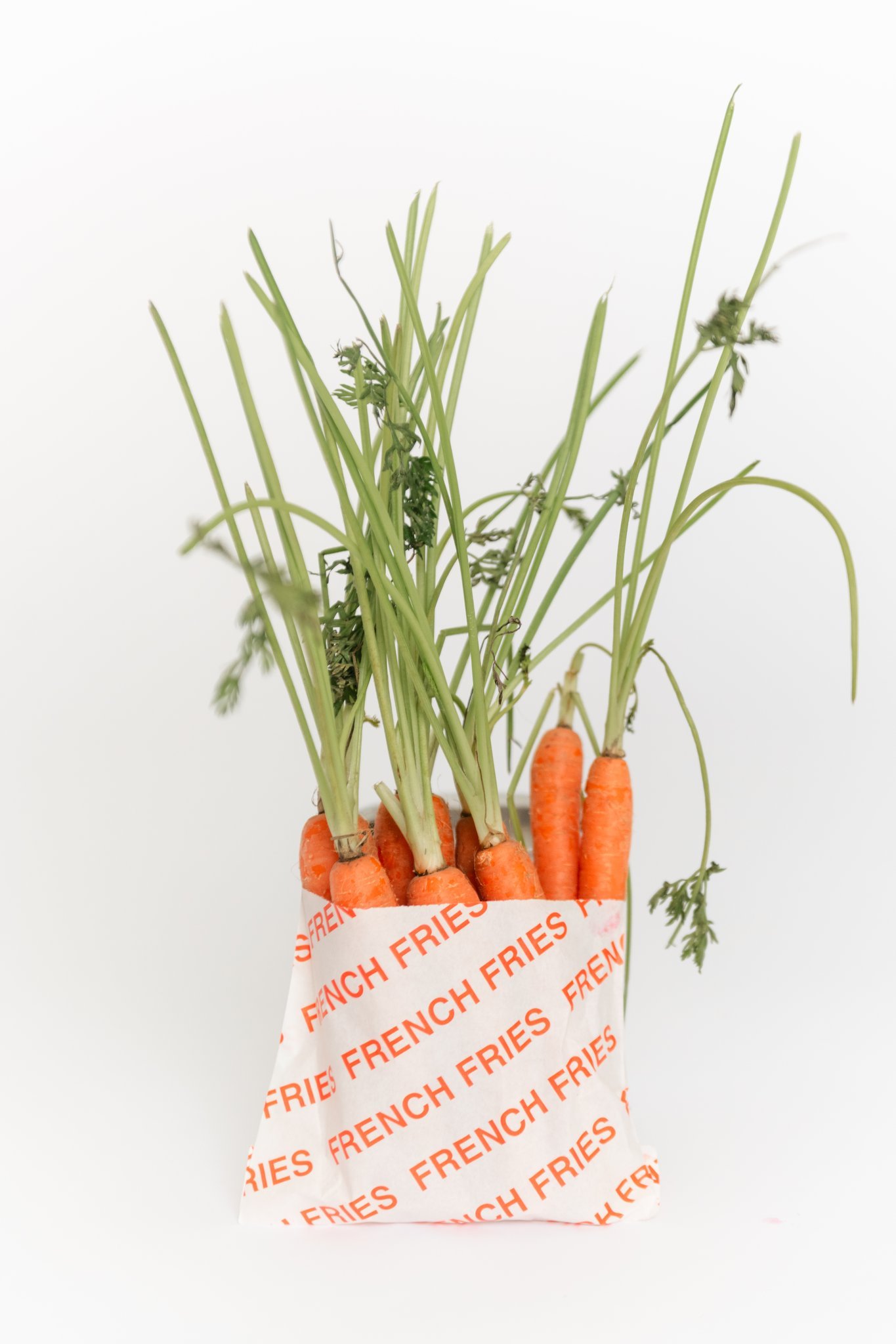 Fresh carrots with green tops in a shopping bag marked "French Fries" on a white background.