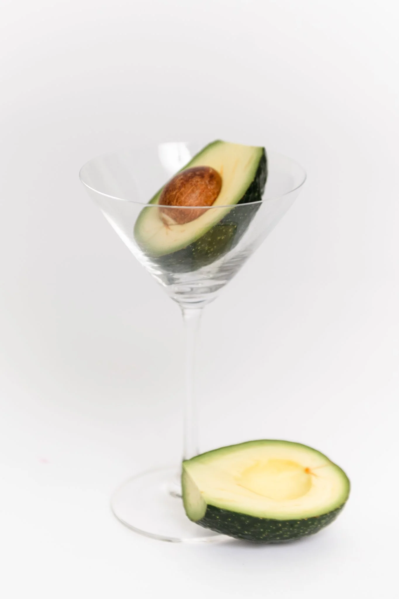 A halved avocado with the seed inside placed on a white surface next to an elegant martini glass containing another piece of avocado.
