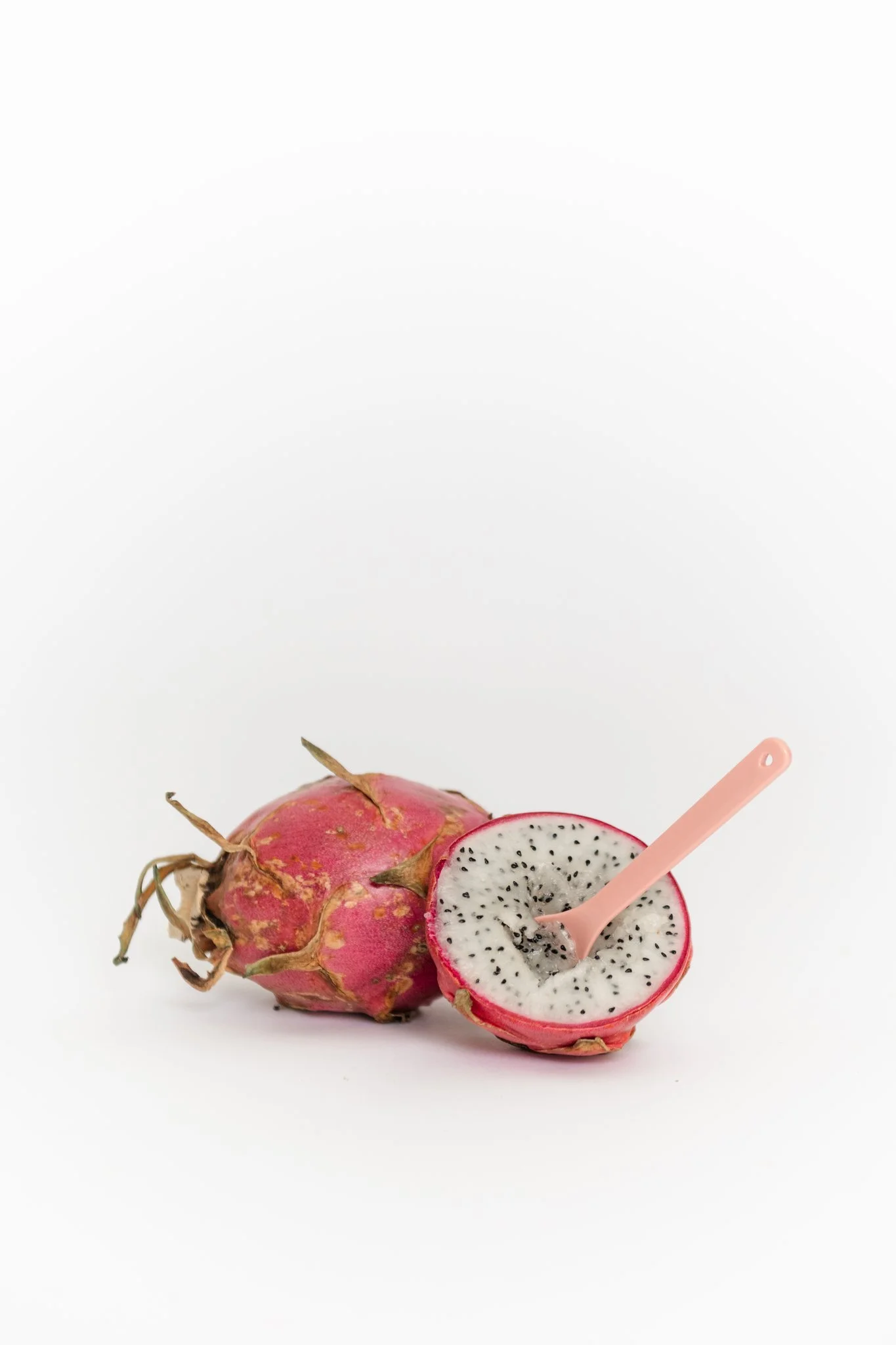 A whole dragon fruit and a half sliced dragon fruit with a pink spoon in the sliced piece, set against a white background.