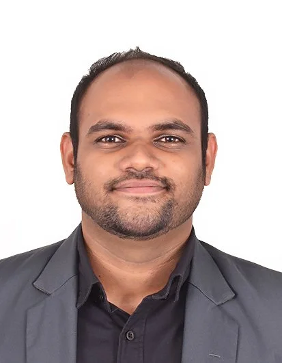 Young man in a black suit, white shirt, black tie, and red-and-white striped medal ribbon hangs around his neck, standing indoors with blurred lights in the background.