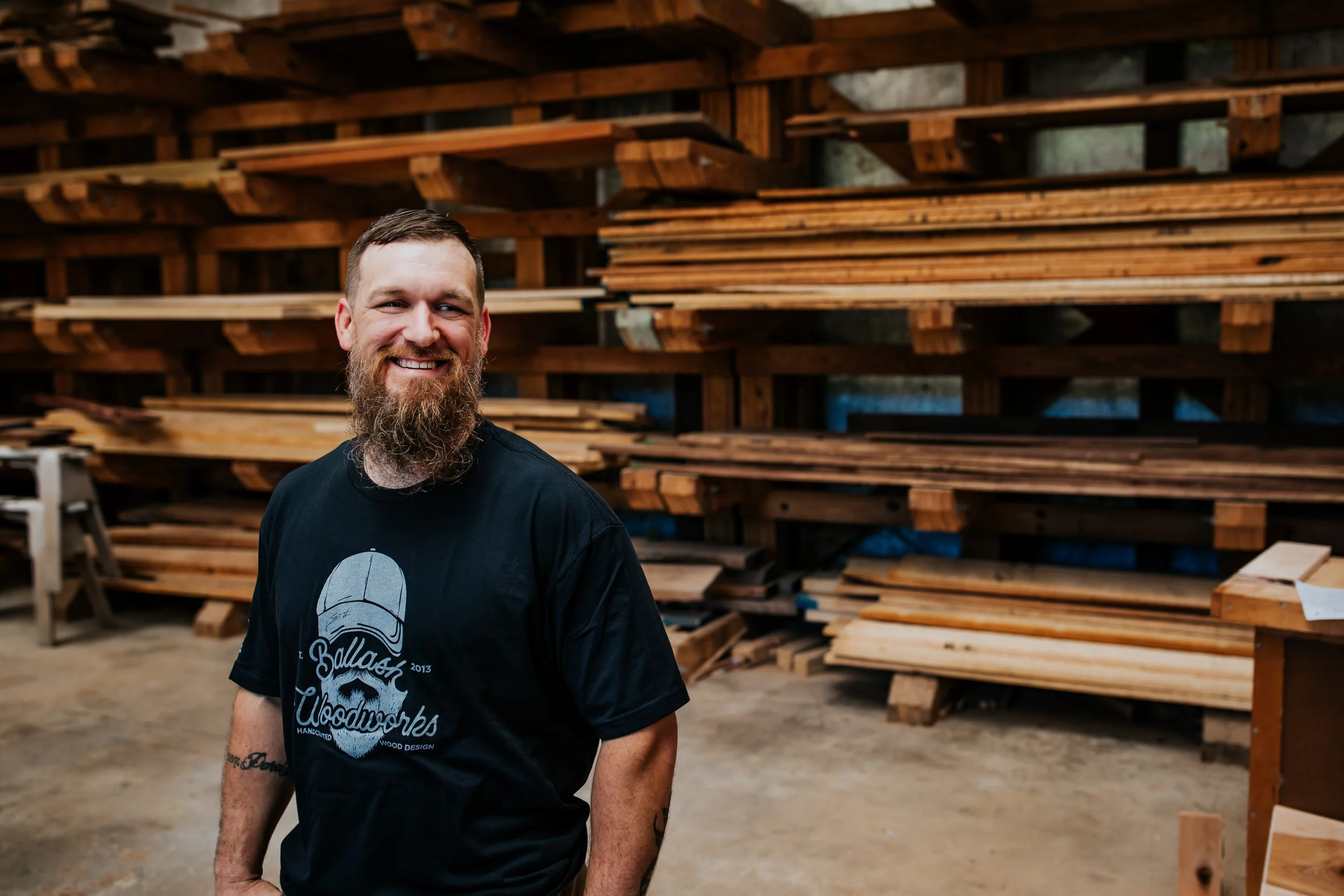 Owner Kurt Ballash standing in front of a wall stacked with wood slabs, looking to the right of the viewer.