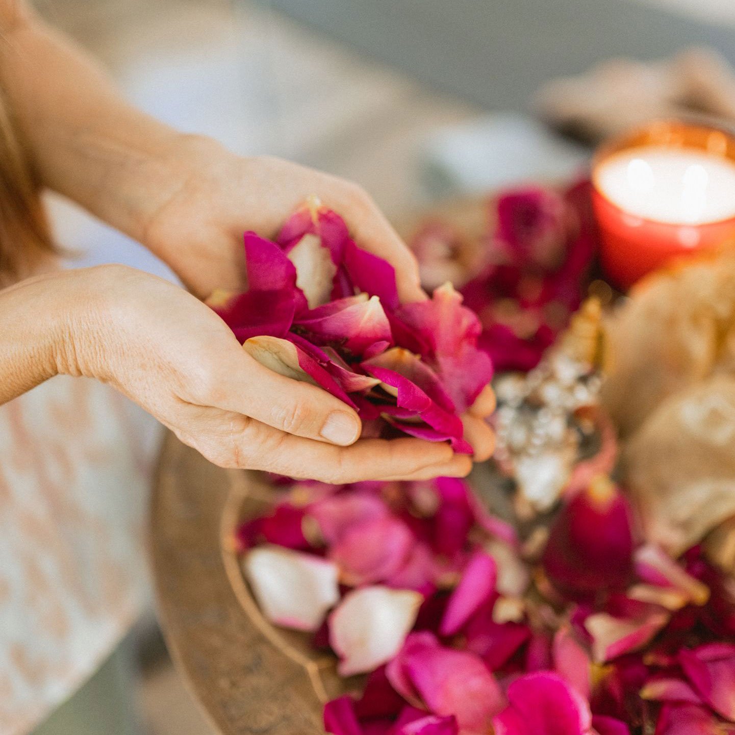 Hand arranging vibrant pink and white rose petals on a table for a ritual or ceremony, with a lit candle in a red holder in the background.