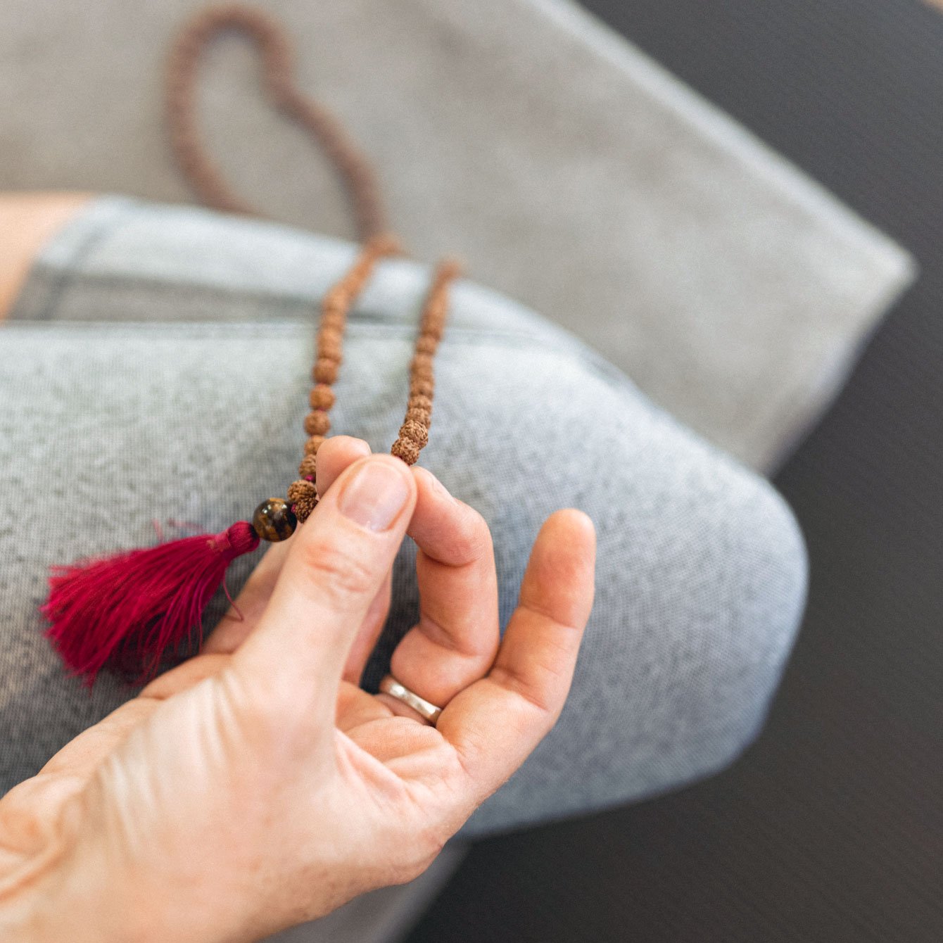 Close-up of a person's hand holding a brown beaded necklace with a red tassel against gray fabric furniture.