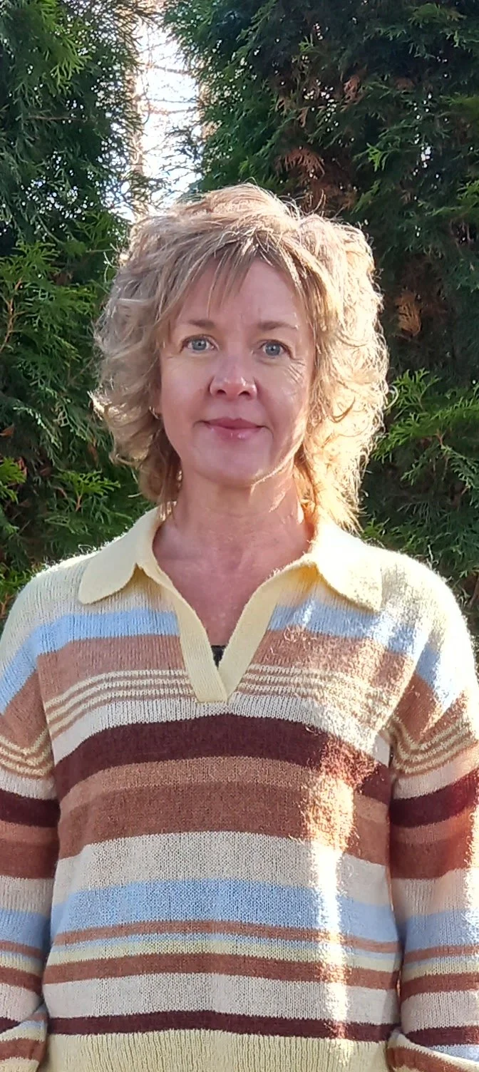 A woman with curly blonde hair and blue eyes is standing outdoors in front of green trees. She is wearing a striped sweater with cream, brown, blue, and maroon colors.