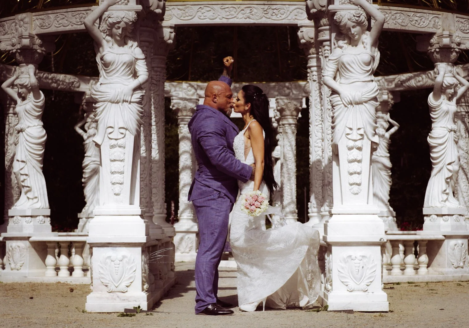 Ein Mann und eine Frau in Hochzeitskleidung, die sich in einer parkähnlichen Umgebung mit weißen Skulpturen und Säulen gegenüberstehen und sich küssen.