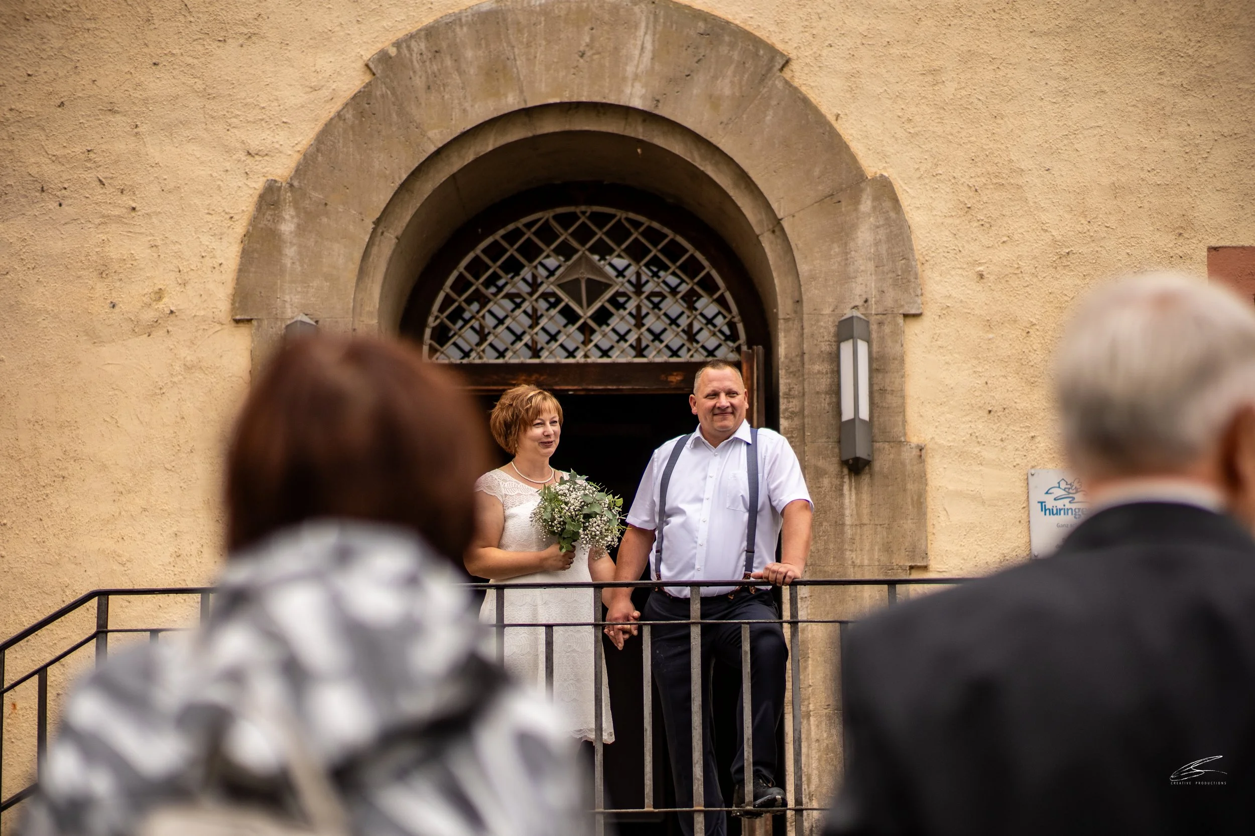 Ein Brautpaar steht auf einer kleinen Plattform vor einem alten Gebäude und hält Händchen. Die Braut trägt ein weißes Kleid und hält einen Blumenstrauß, der Bräutigam trägt ein weißes Hemd mit Hosenträgern. Einige Zuschauer sind im Vordergrund zu seh