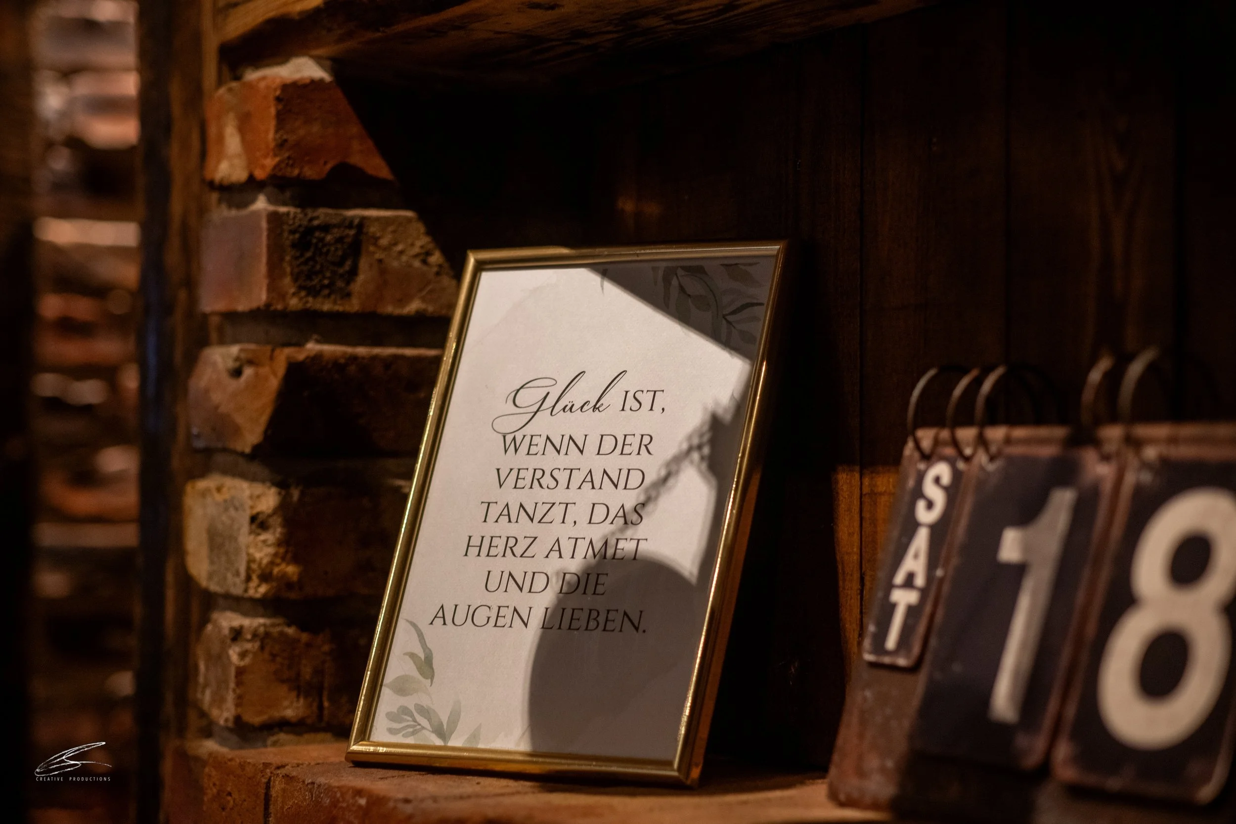 Ein gerahmtes Zitat auf einem Holzregal, das auf einer Ziegelwand steht. Das Zitat lautet: ‚Glück ist, wenn der Verstand tanzt, das Herz atmet und die Augen lieben.‘ Neben dem Rahmen sind Digitalanzeigen mit den Nummern 18.
