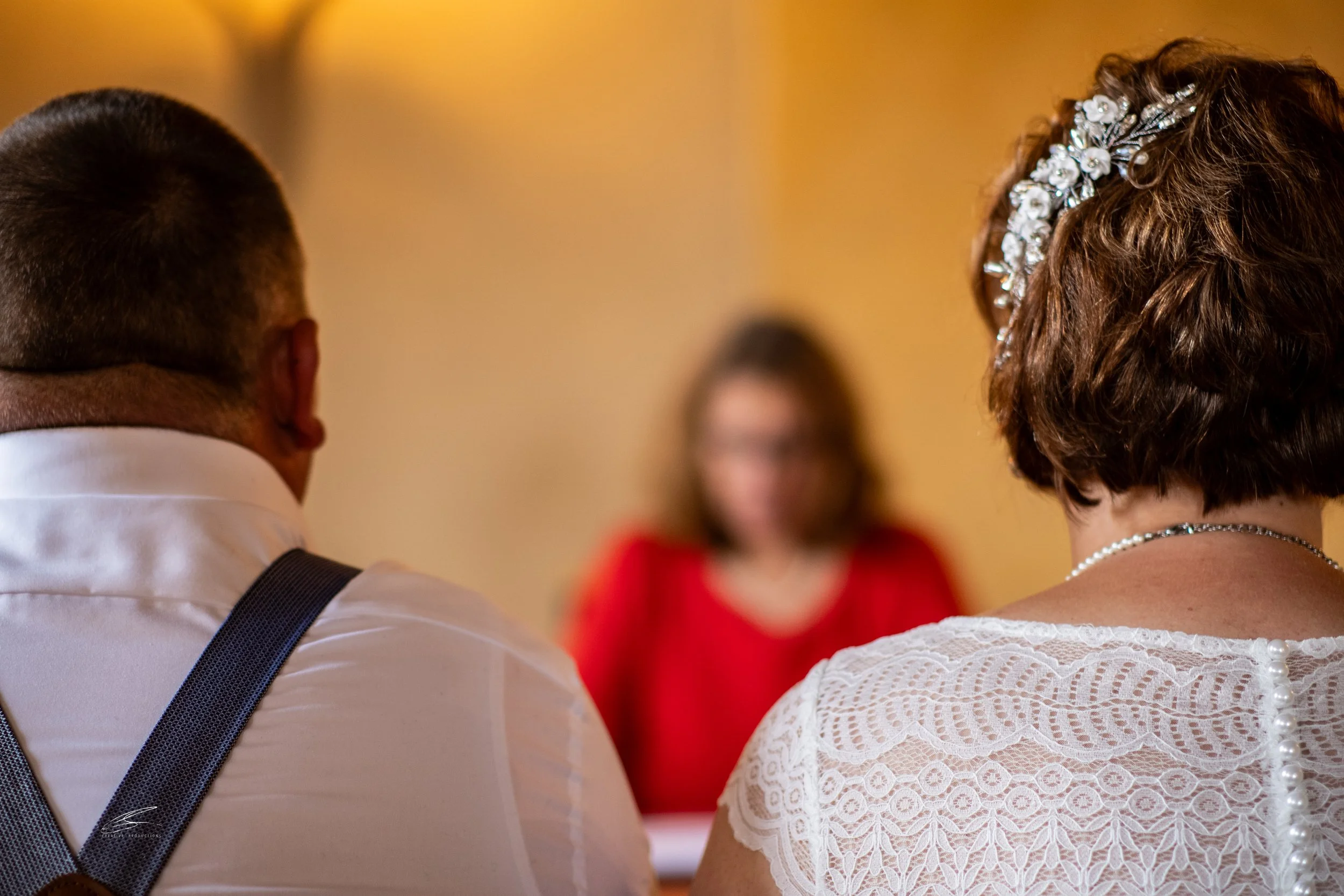 Nahaufnahme von einem Brautpaar, das auf ihre Zeremonie wartet, mit einer Frau im Hintergrund, die bei der Hochzeitszeremonie spricht.