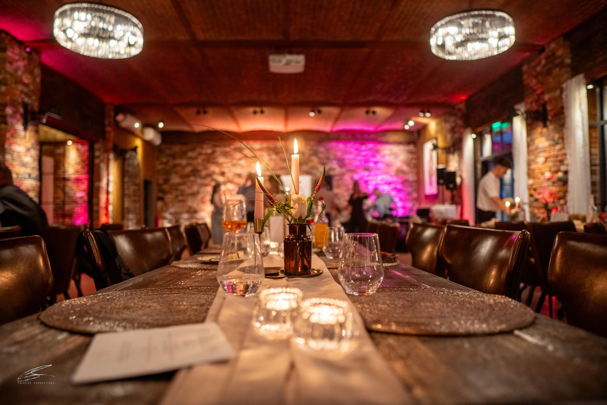 Ein festlich dekorierter Esstisch mit Kerzen, Gläsern, Tellern und Blumen in einem Restaurant mit rotem Ziegelsteinwänden und warmem Ambiente