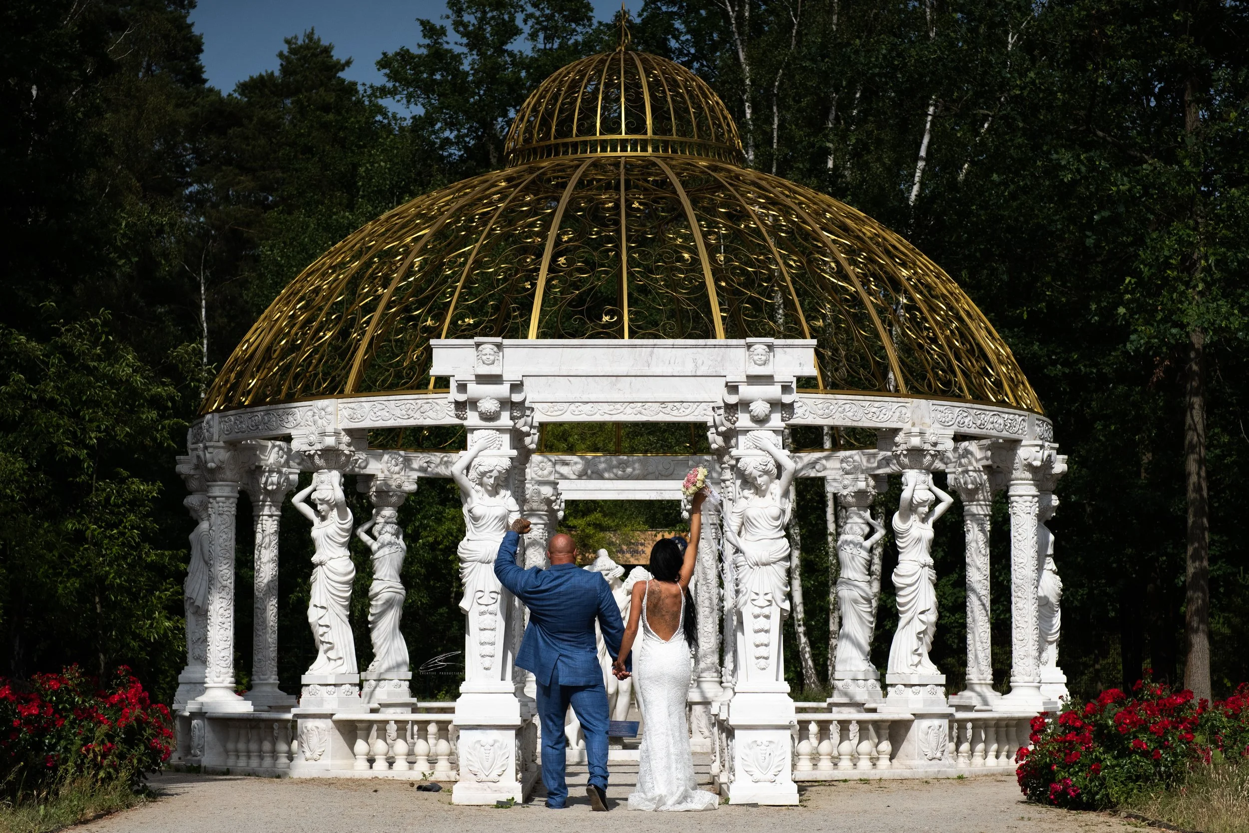 Ein Brautpaar hält sich an den Händen, während sie sich einer eleganten weißen Pavillon mit goldener Kuppel nähern, umgeben von grünen Bäumen und roten Blumen.