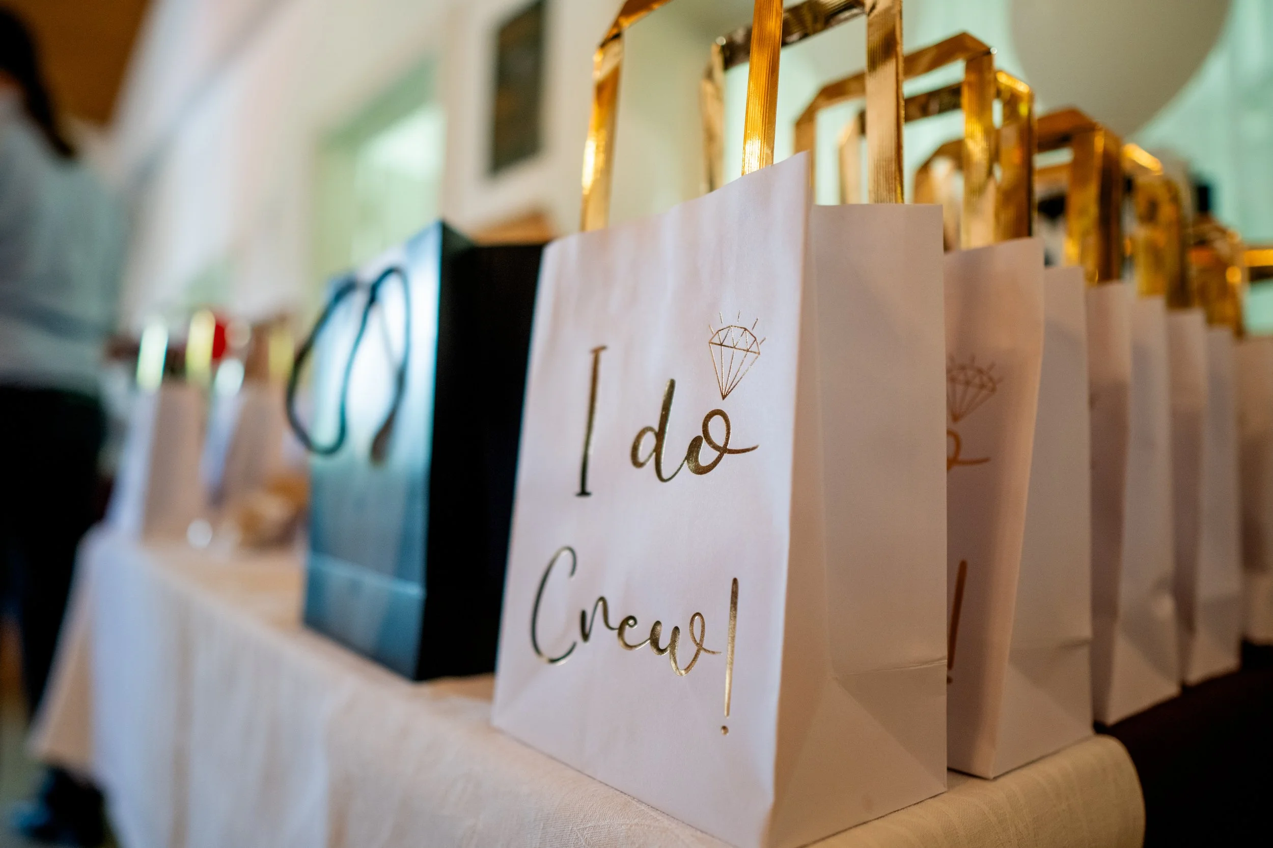 Geschenkstüten, eine davon mit der Aufschrift 'I do Crew' in Goldschrift und einem Diamant-Logo, auf einem Tisch bei einer Veranstaltung.