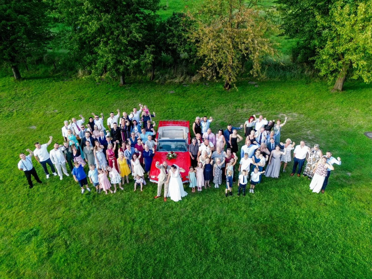Hochzeit mit einer großen Gruppe von Menschen im Grünen, in der Mitte ein rotes Auto, um das sich die Gäste versammeln, einige feiern und posieren für die Kamera.