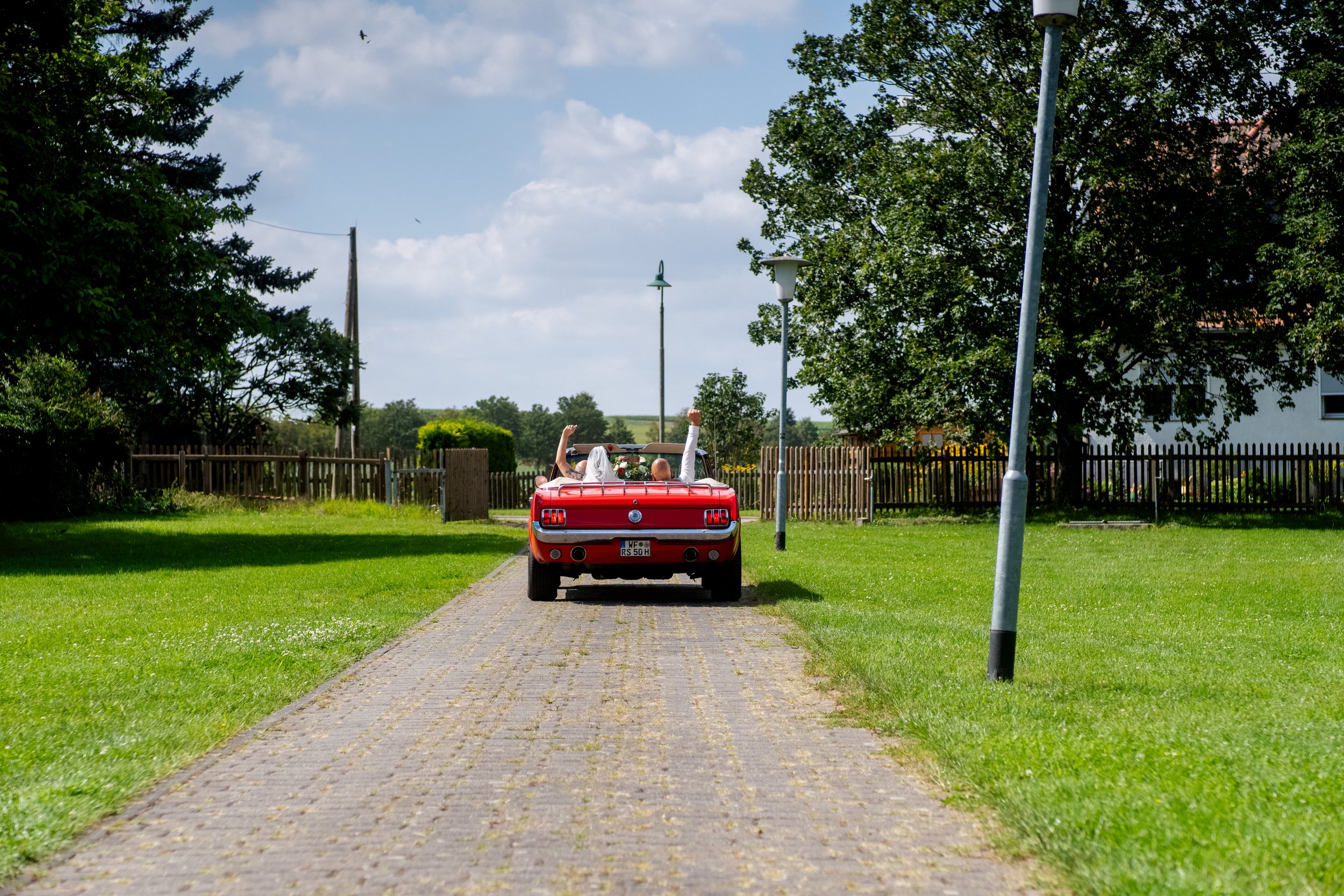 Zwei Eheleute sitzen in einem roten Ford Mustang Cabrio, das auf einer gepflasterten Straße in einem grünen Park parkt, mit Blick auf den blauen Himmel und Bäume im Hintergrund.