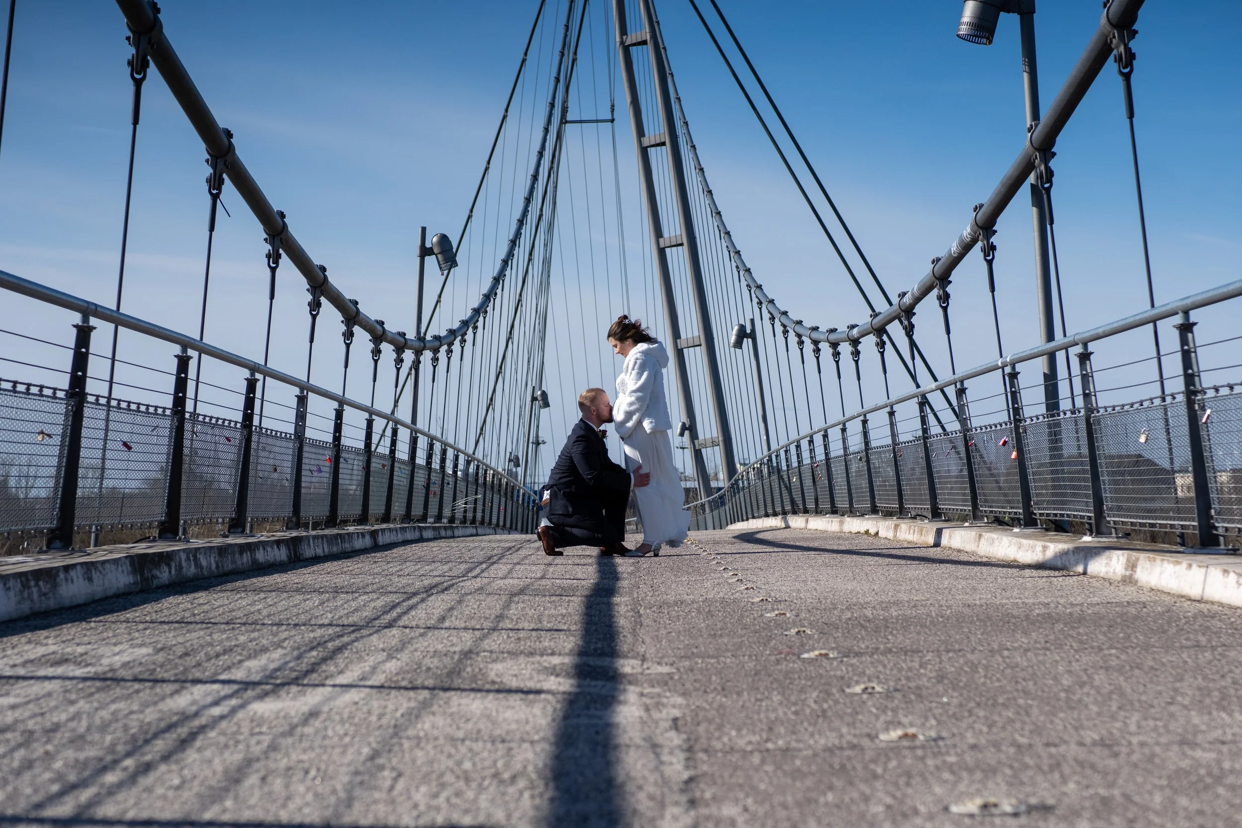 Ein Brautpaar auf einer Brücke bei Sonnenaufgang oder Sonnenuntergang, die Frau in einem weißen Hochzeitskleid und der Mann in einem dunklen Anzug, während er auf den Knien vor ihr ist.
