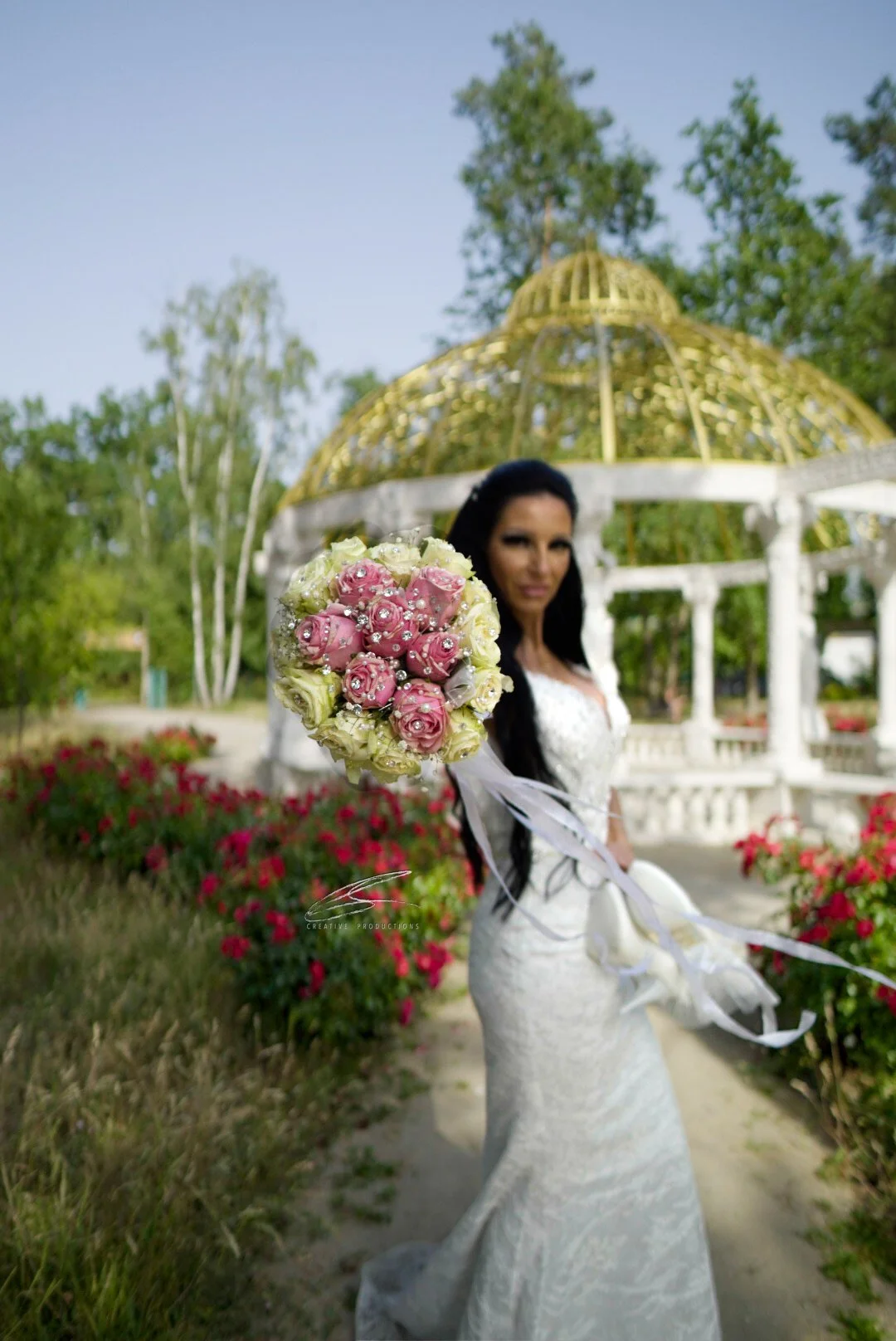 Eine Frau in einem weißen Hochzeitskleid hält einen Blumenstrauß aus rosa und weißen Rosen in der Hand. Im Hintergrund befindet sich ein Pavillon mit goldener Kuppel, umgeben von einem Garten mit Blumen und Bäumen.