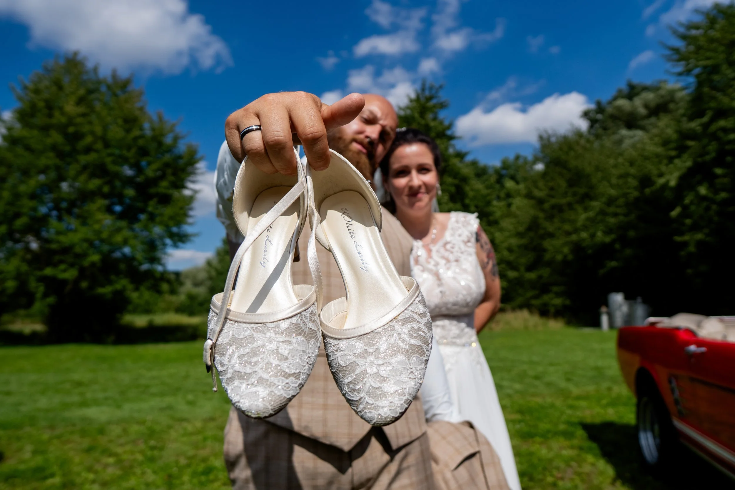 Ein Mann hält ein Paar weiße, spitz zulaufende Brautschuhen vor der Kamera, während eine Frau im weißen Brautkleid im Hintergrund auf einer grünen Wiese bei sonnigem Himmel steht.