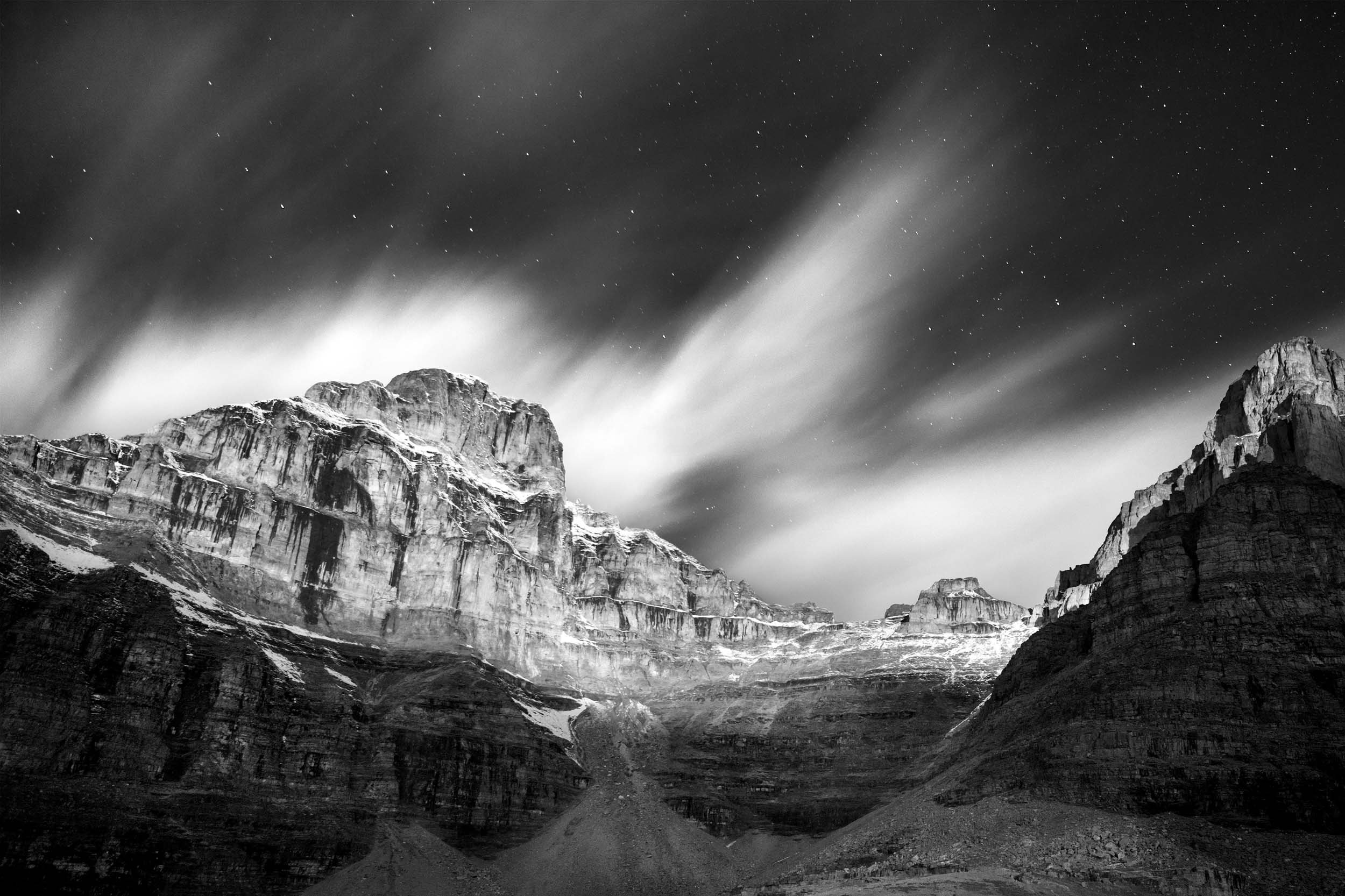 Snowy mountain lit by moonlight with wispy clouds and stars in the night sky.