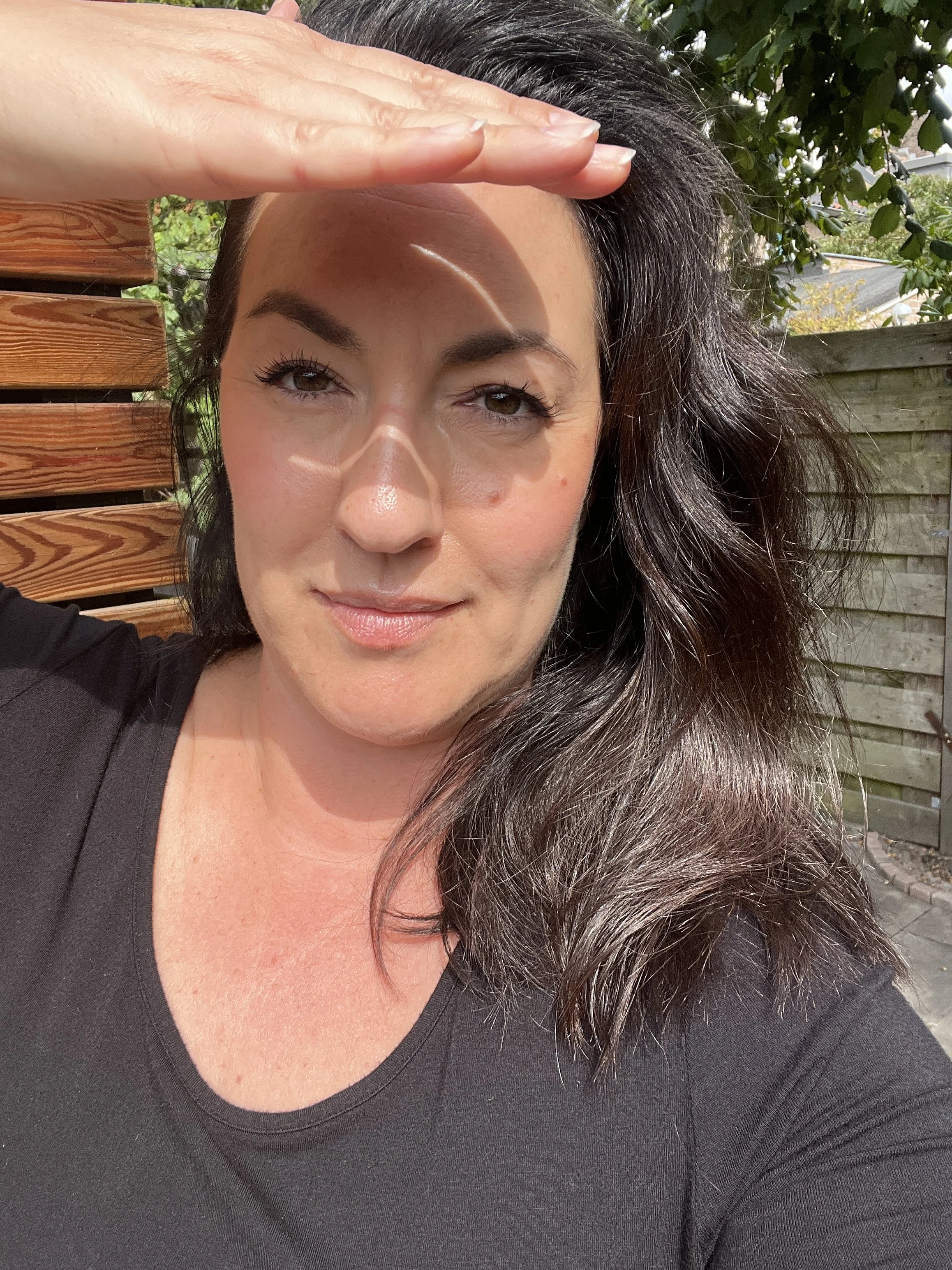 A woman with long dark hair, wearing a black top, is outdoors shielding her eyes from the sun with her hand. She has light brown eyes and makeup, and is standing in front of a wooden fence with greenery behind her.